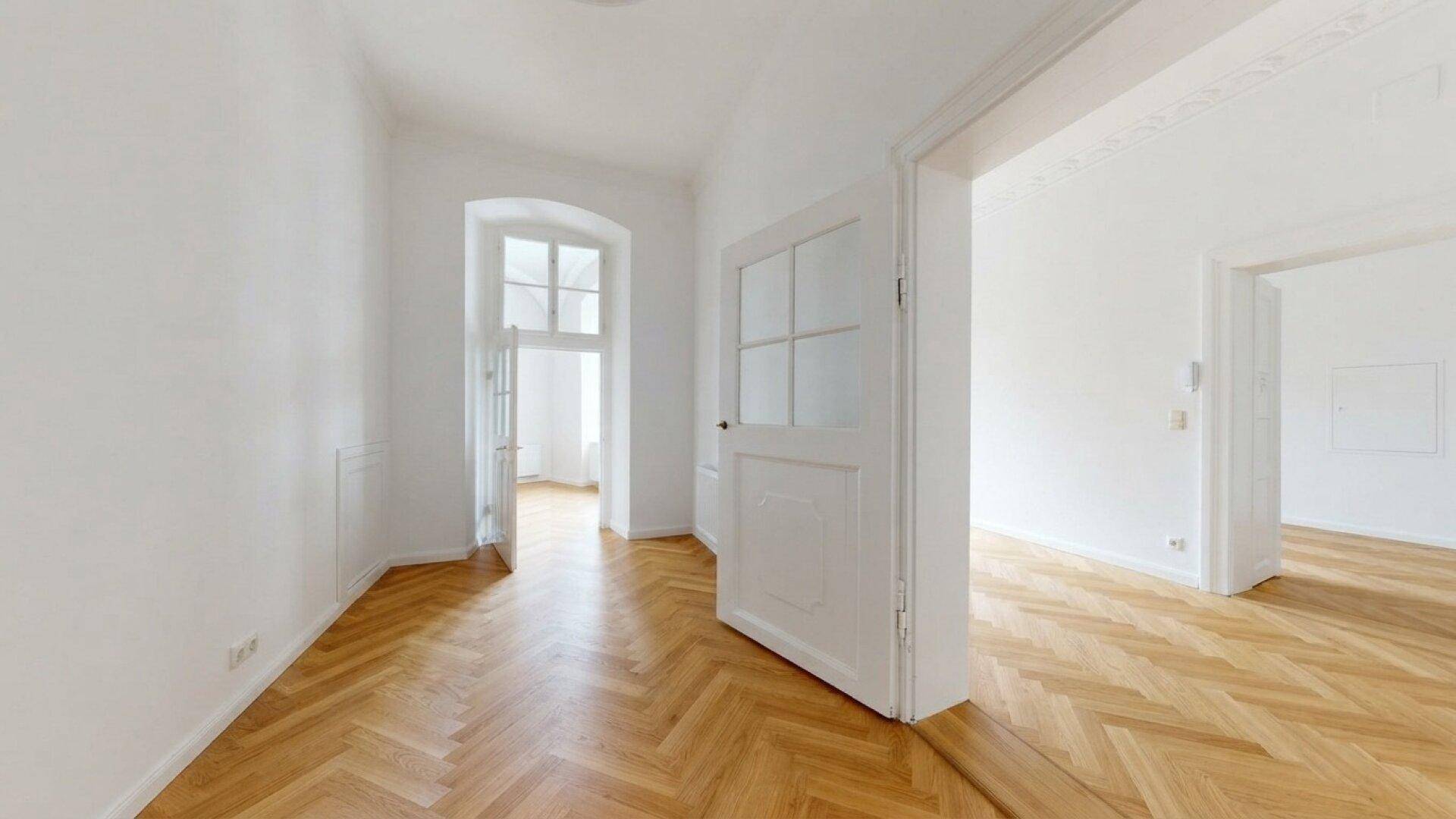 Heller Flur mit Fischgrätparkettboden, weißen Wänden und einem Bogenfenster am Ende.