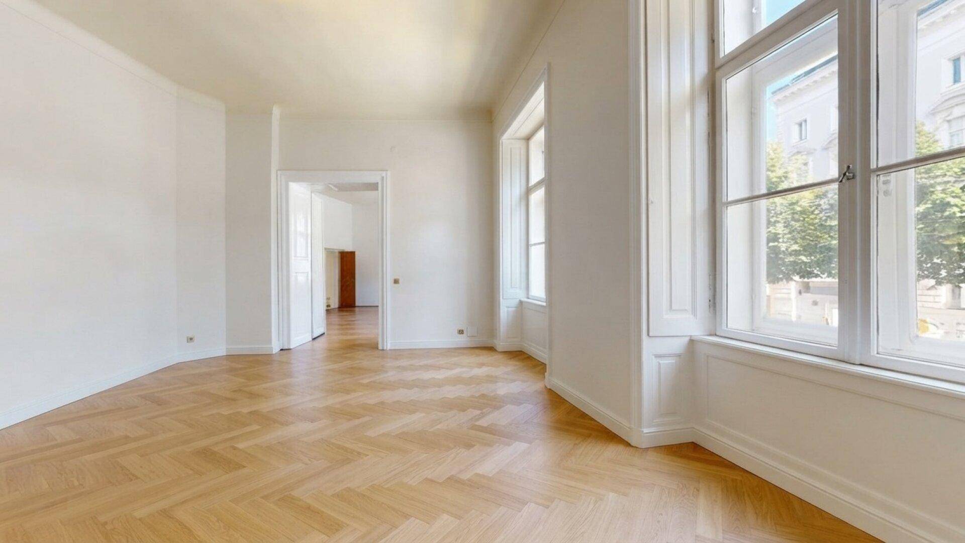Heller, leerer Raum mit hohen Fenstern, Fischgrätparkett und Zugang zu weiteren Zimmern.