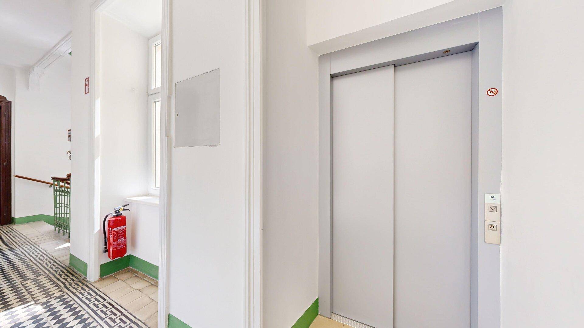 Heller Flur im Gebäude mit einem modernen Aufzug und einem Feuerlöscher an der Wand.