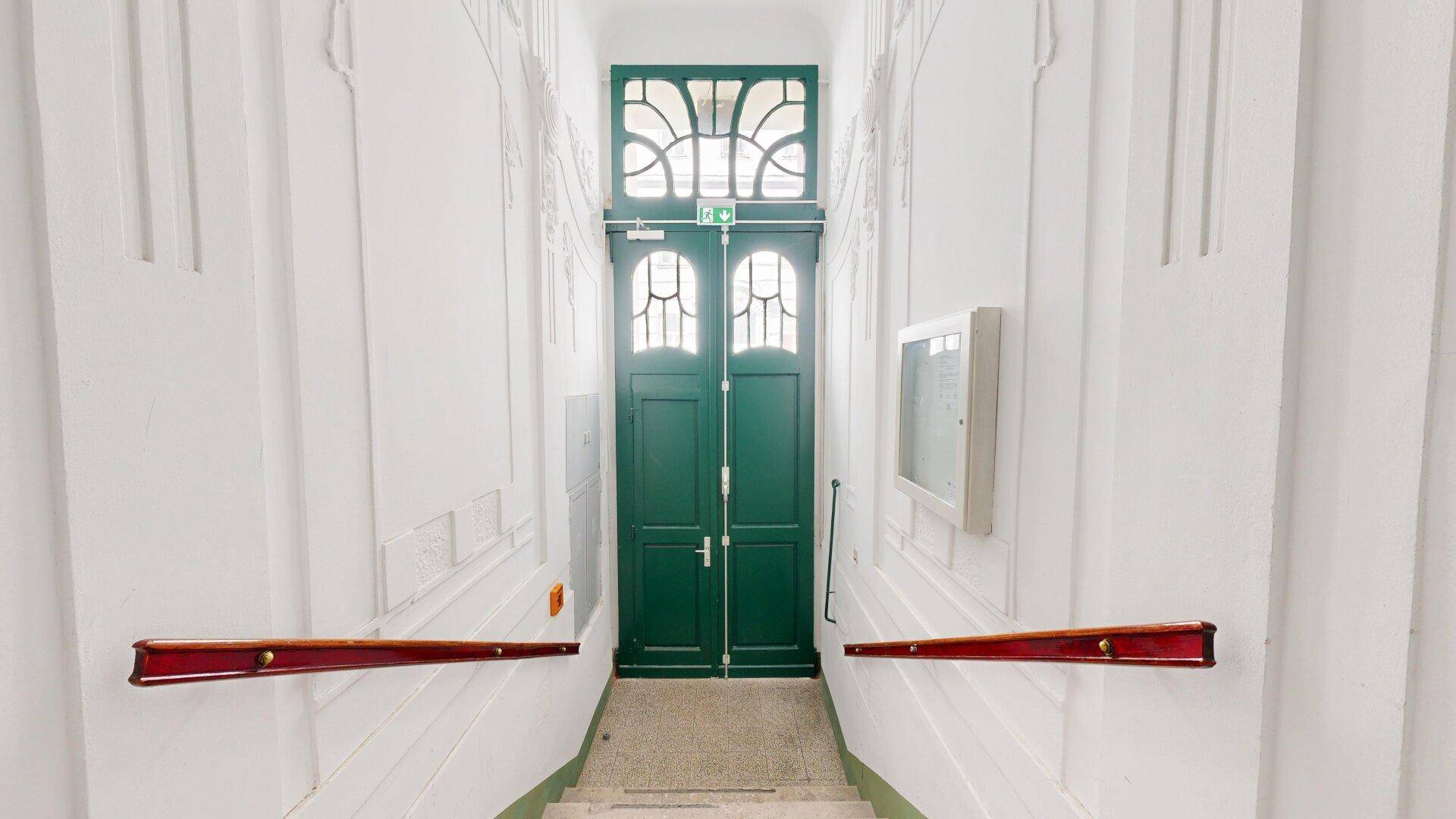 Treppenhaus mit verzierten Handläufen und einer großen, grünen Doppeltür am Ende des Ganges.