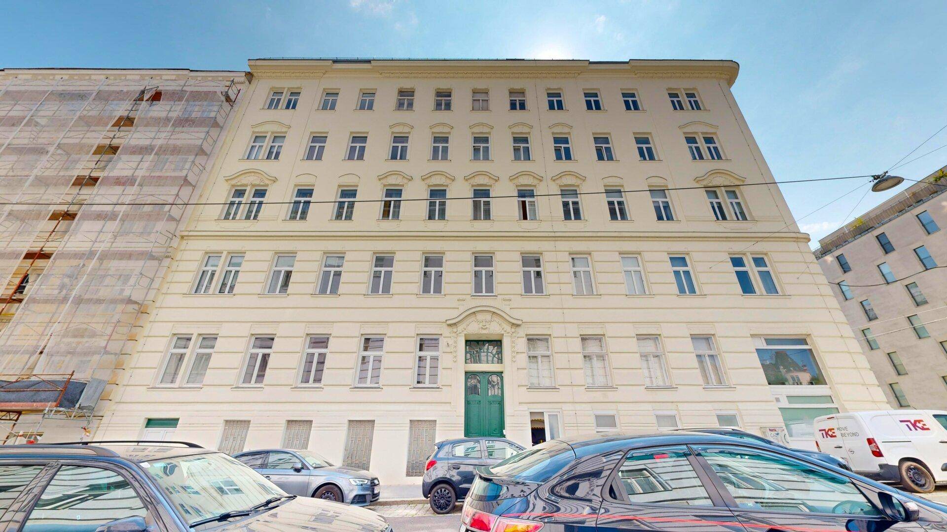 Klassisches Mehrfamilienhaus mit vielen Fenstern und einer grünen Eingangstür an einer belebten Straße.
