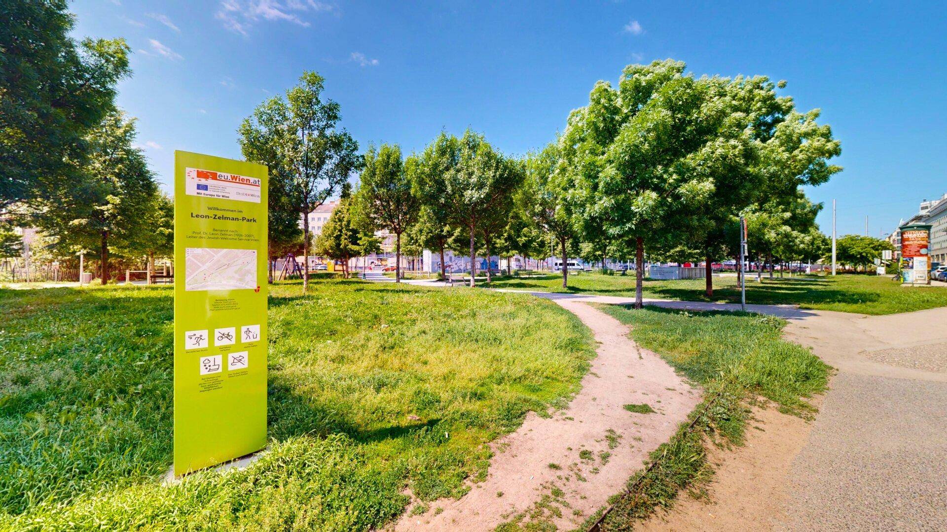 Grüner Stadtpark mit Bäumen, einem Spazierweg und einer Informationstafel unter blauem Himmel.