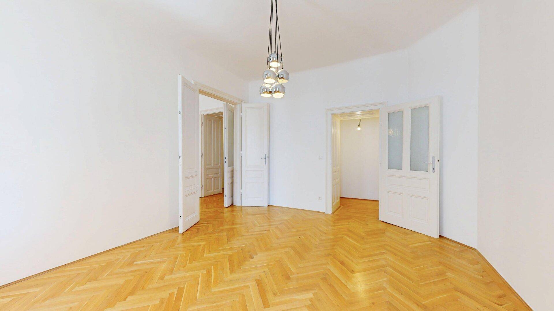 Geräumiger Raum mit Fischgrätparkett und mehreren Türen, die verschiedene Bereiche der Wohnung verbinden.
