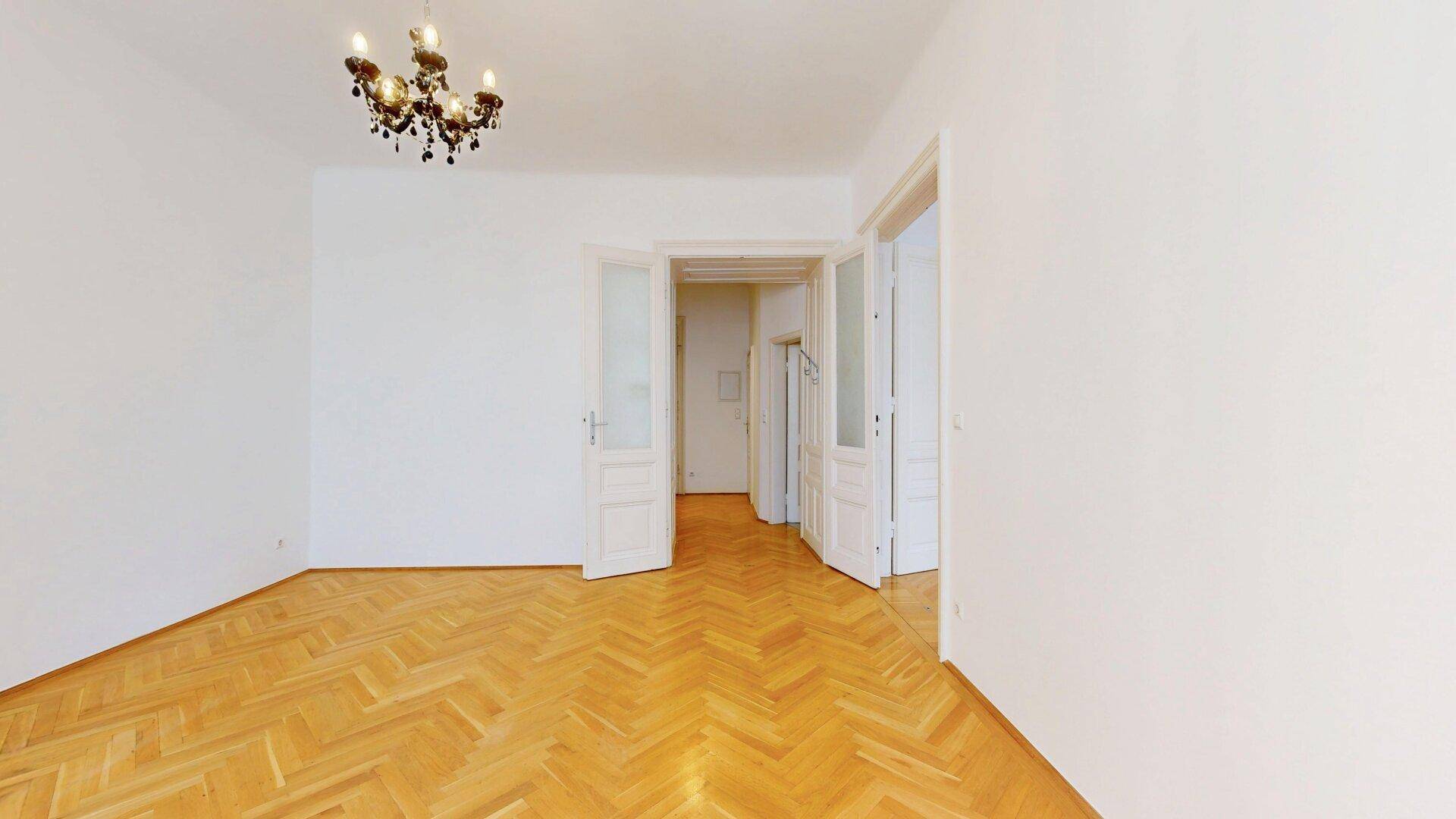Leeres Wohnzimmer mit Fischgrätparkett und zwei Flügeltüren, die zu weiteren Räumen führen.