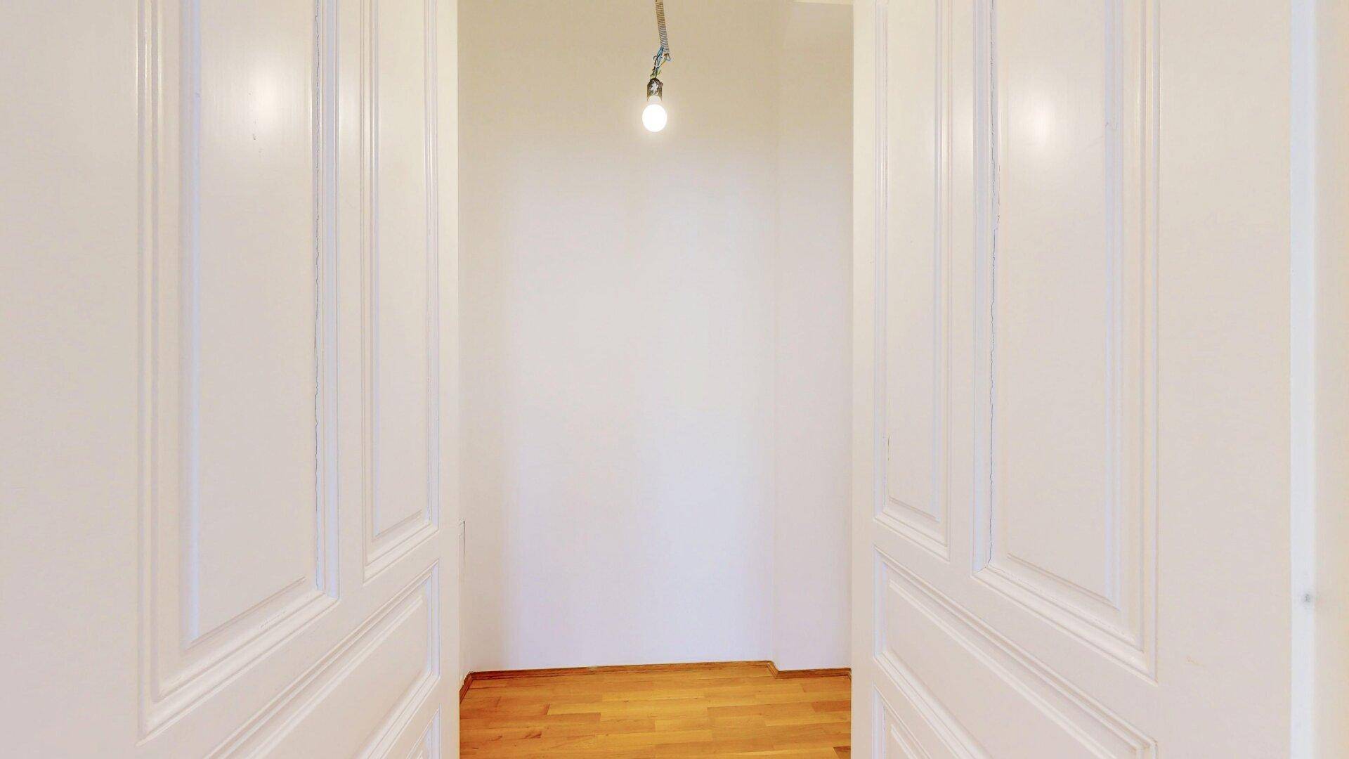 Blick durch eine offene Doppeltür in einen leeren Flur mit einfacher Glühbirnenbeleuchtung.