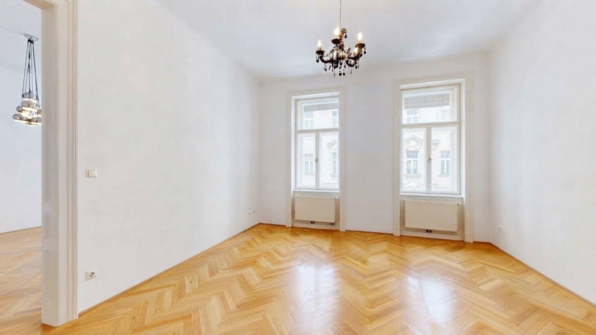 Geräumiges Wohnzimmer mit klassischem Kronleuchter, Fischgrätparkett und zwei großen Fenstern.
