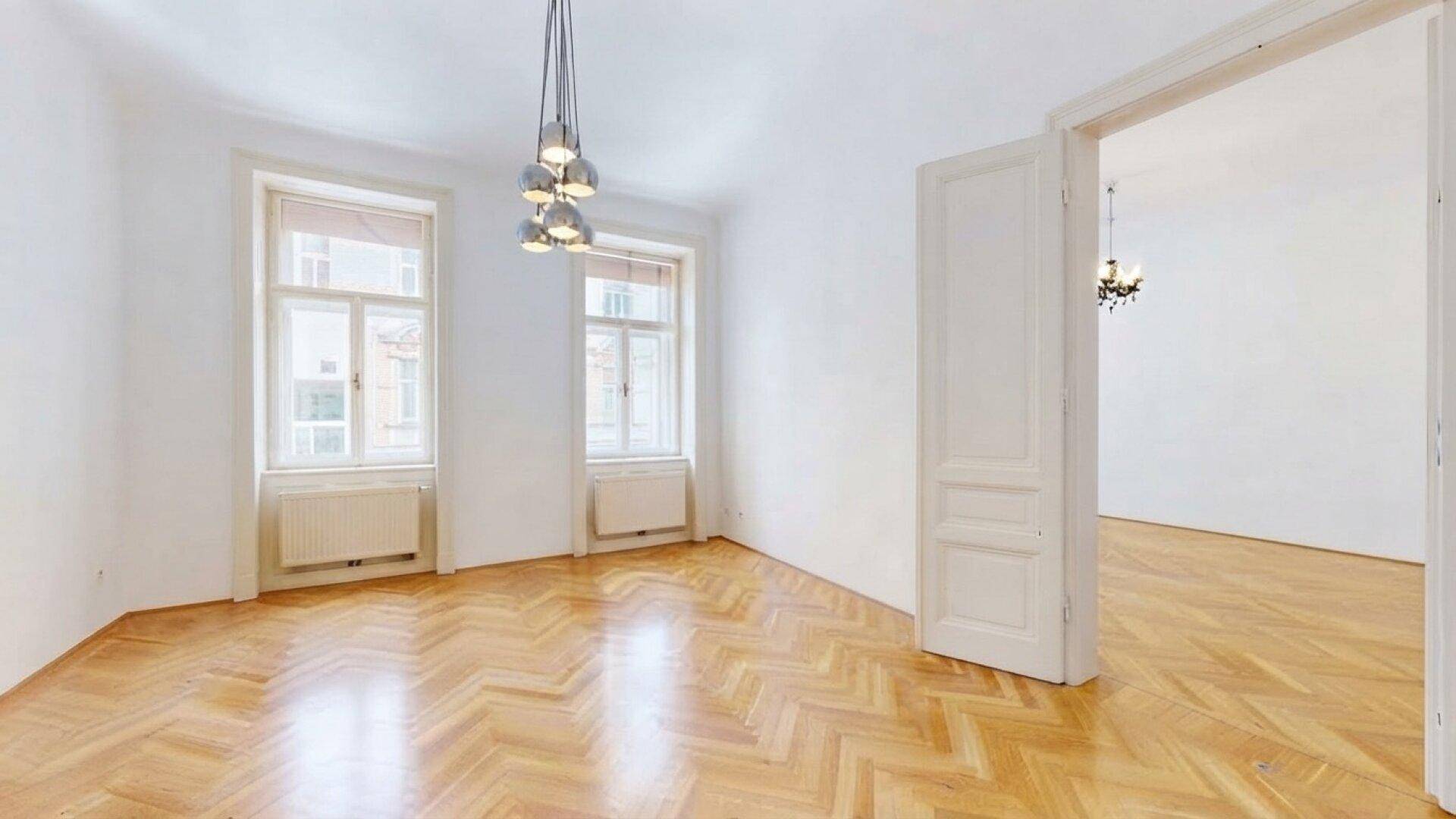 Heller Wohnraum mit Fischgrätparkett, zwei Fenstern und einer offenen Tür zu einem angrenzenden Zimmer.