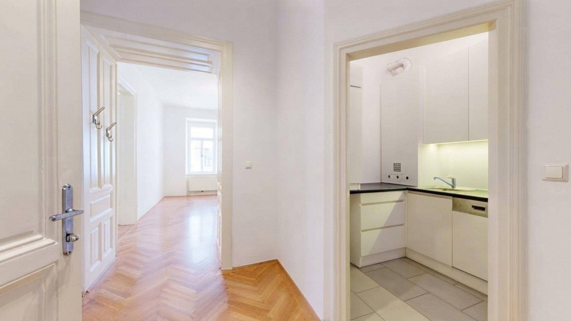 Flur mit Fischgrätparkett, der zu einer kleinen, funktionalen Küche und weiteren Räumen führt.