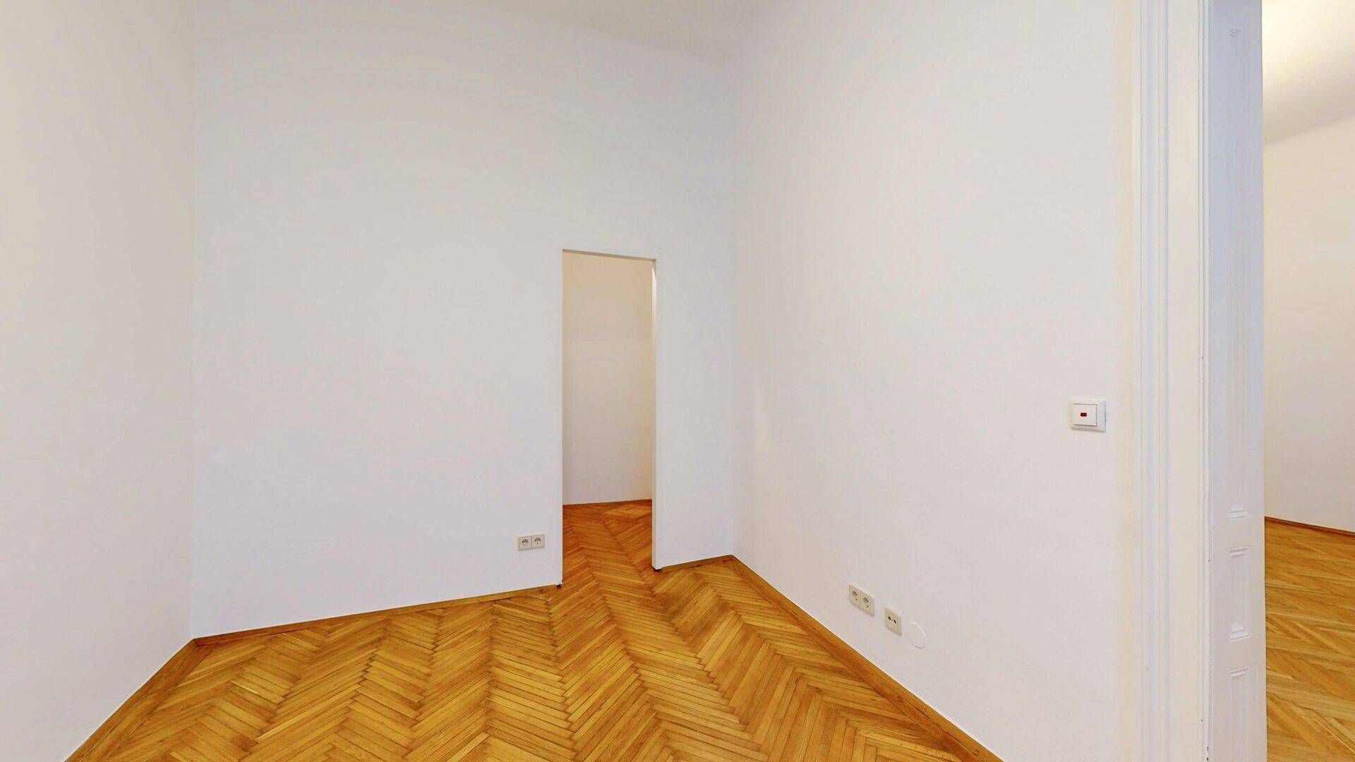 Leeres Schlafzimmer mit Fischgrätparkett und einem offenen Durchgang zu einem weiteren Raum.