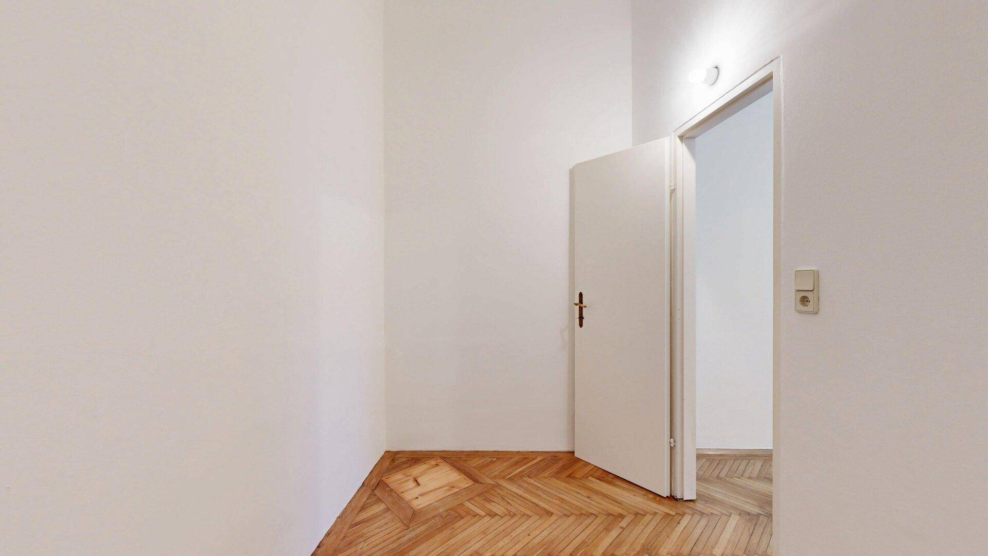 Einfaches Schlafzimmer mit Fischgrätparkett und einer geöffneten Tür, die den Blick in einen weiteren Raum freigibt.