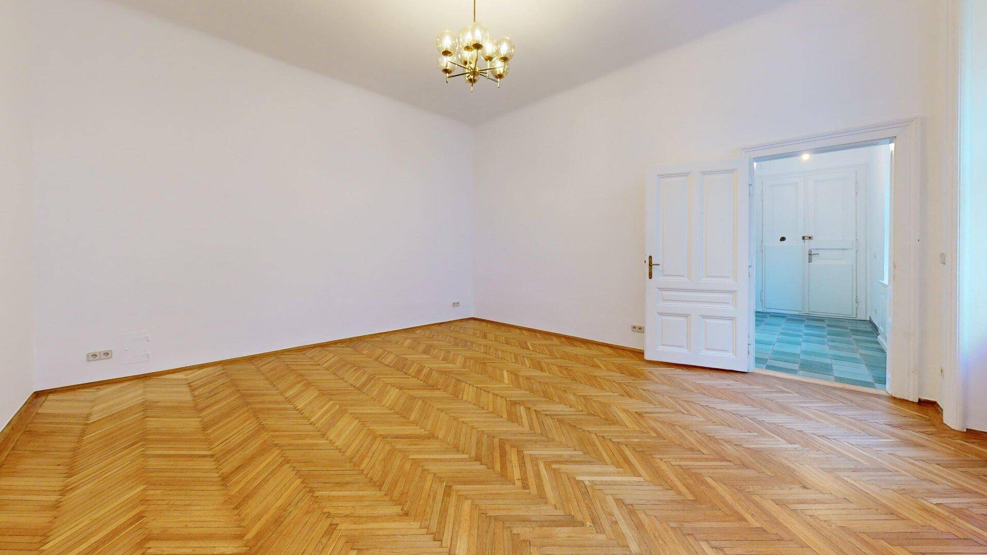 Eckansicht eines leeren Wohnzimmers mit glänzendem Fischgrätparkett und einer Tür, die zu einem gefliesten Bereich führt.
