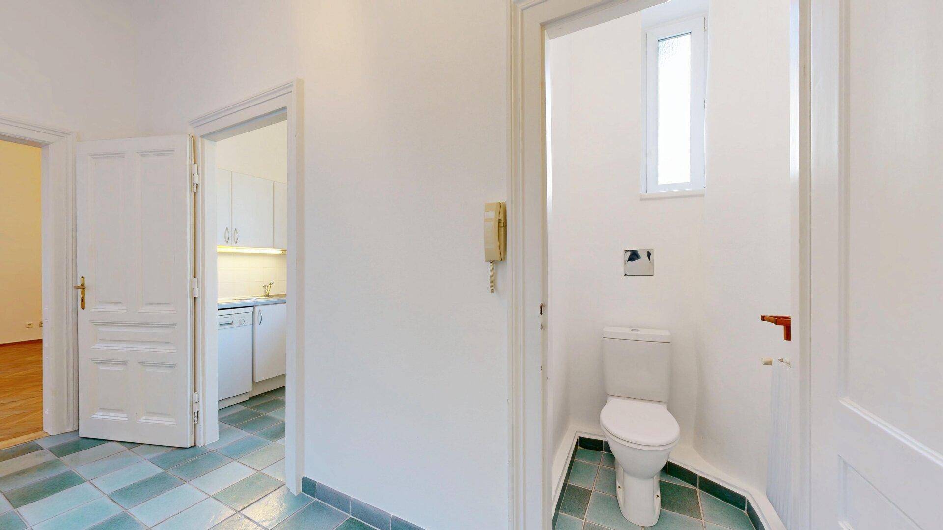 Separates WC mit Fenster, weißer Toilette und grün-weiß karierten Fliesen, neben dem Eingangsbereich.