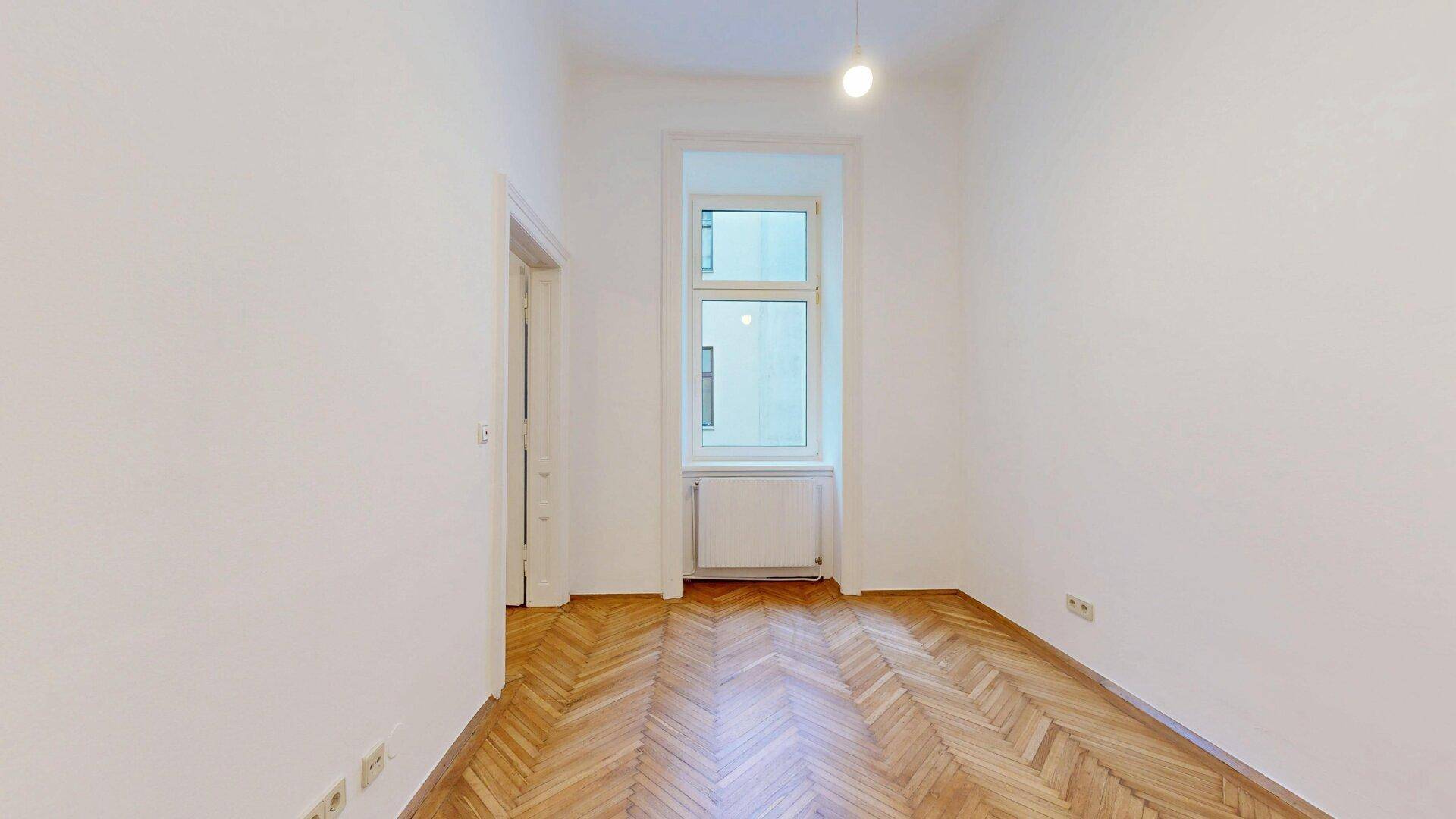 Helles Schlafzimmer mit Fischgrätparkett, einem großen Fenster und einem Heizkörper darunter.