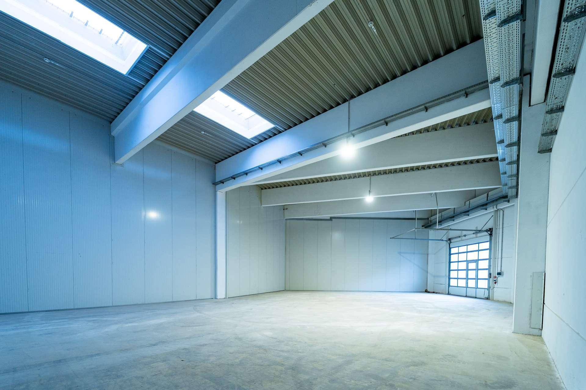 Geräumige, helle Lagerhalle mit hohen Decken, Betonboden und Oberlichtern für natürliches Licht.