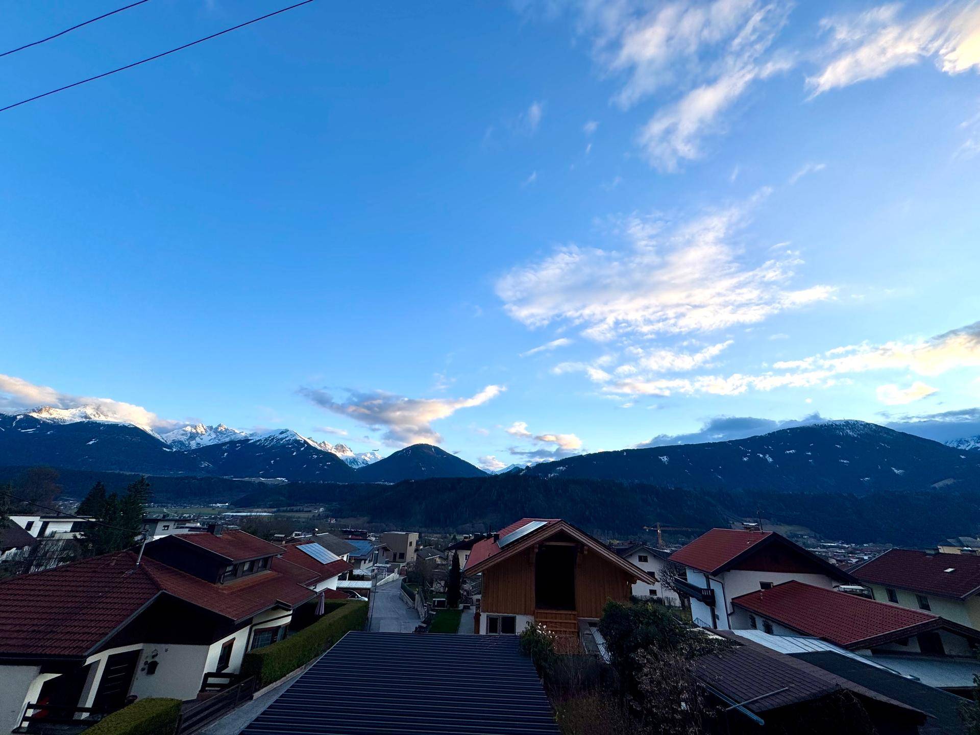 Panoramablick über eine Wohnsiedlung mit roten Dächern und majestätischen Bergen im Hintergrund unter blauem Himmel.