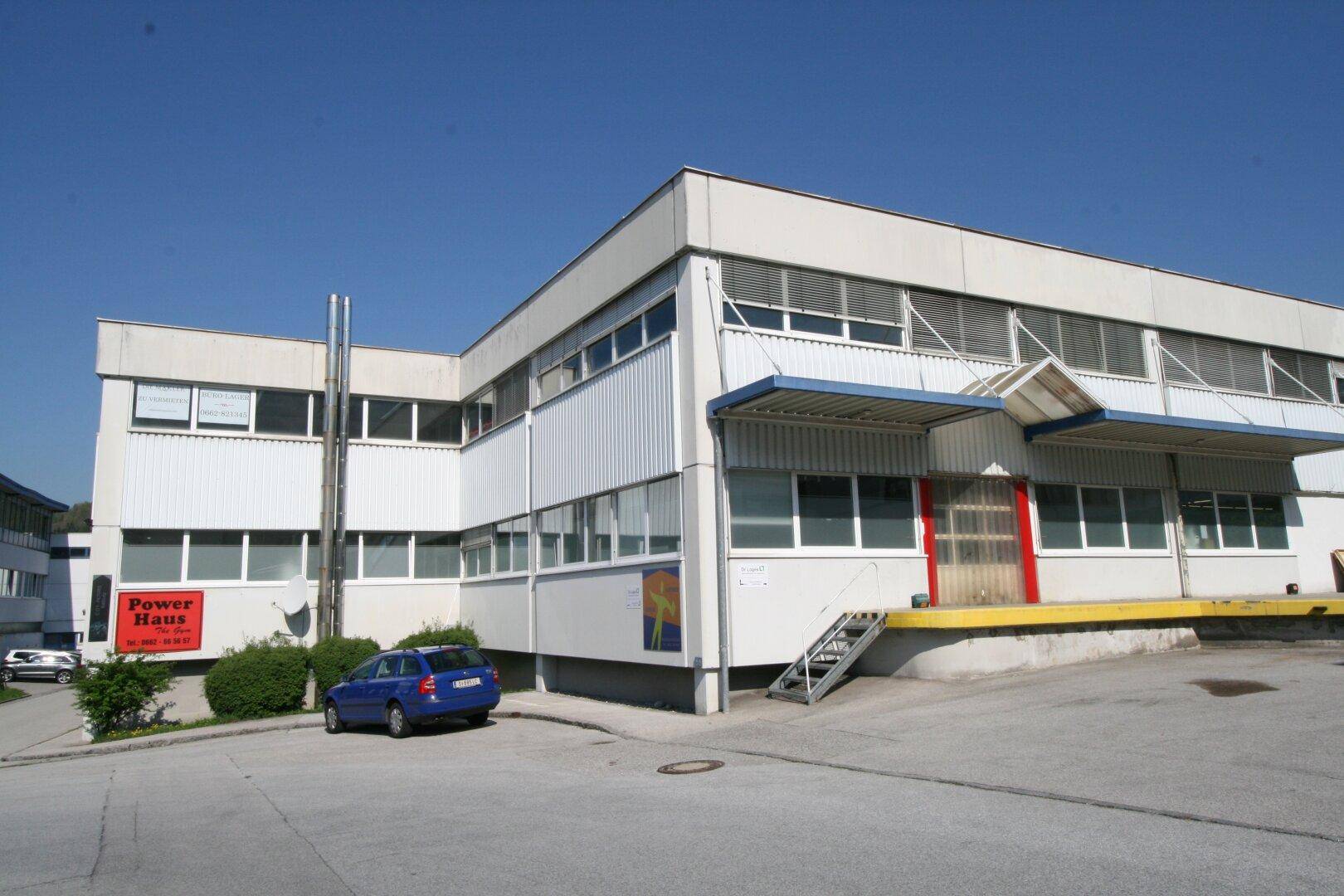 Gewerbegebäude mit Laderampe, Parkplätzen und moderner Architektur, umgeben von grüner Landschaft.