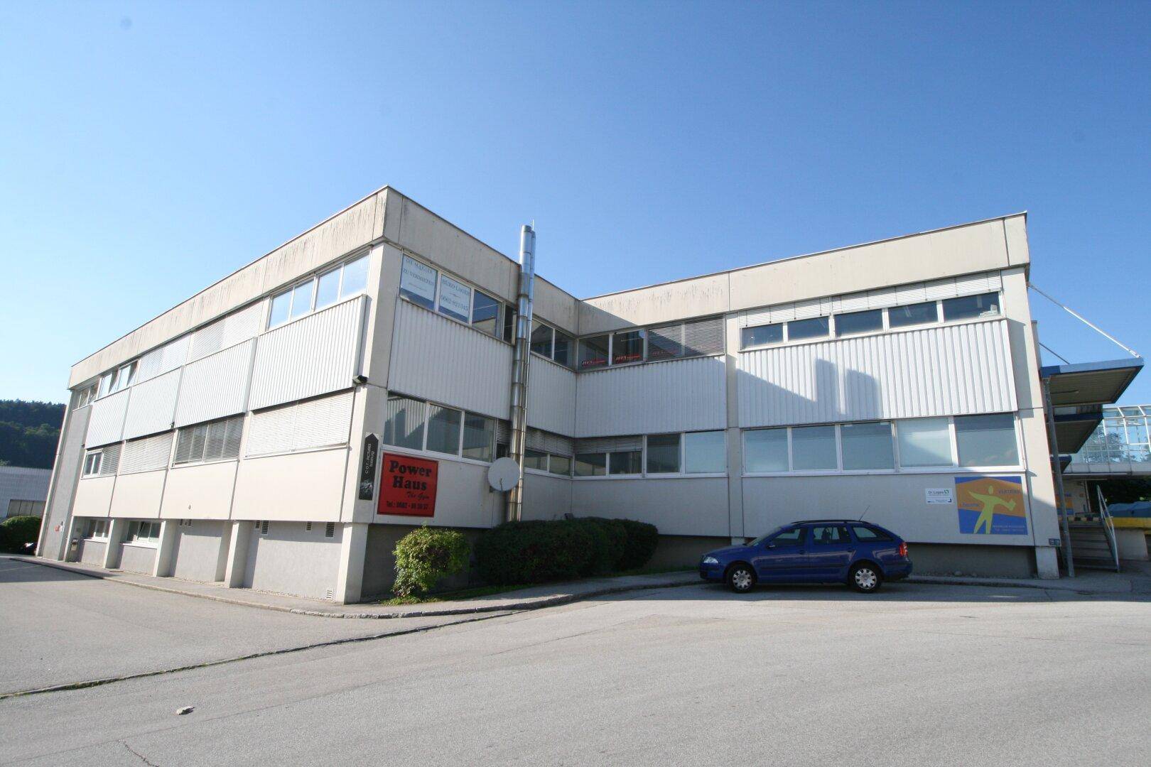 Modernes Geschäftsgebäude mit mehreren Etagen, großen Fenstern und einem Parkplatz im Freien.