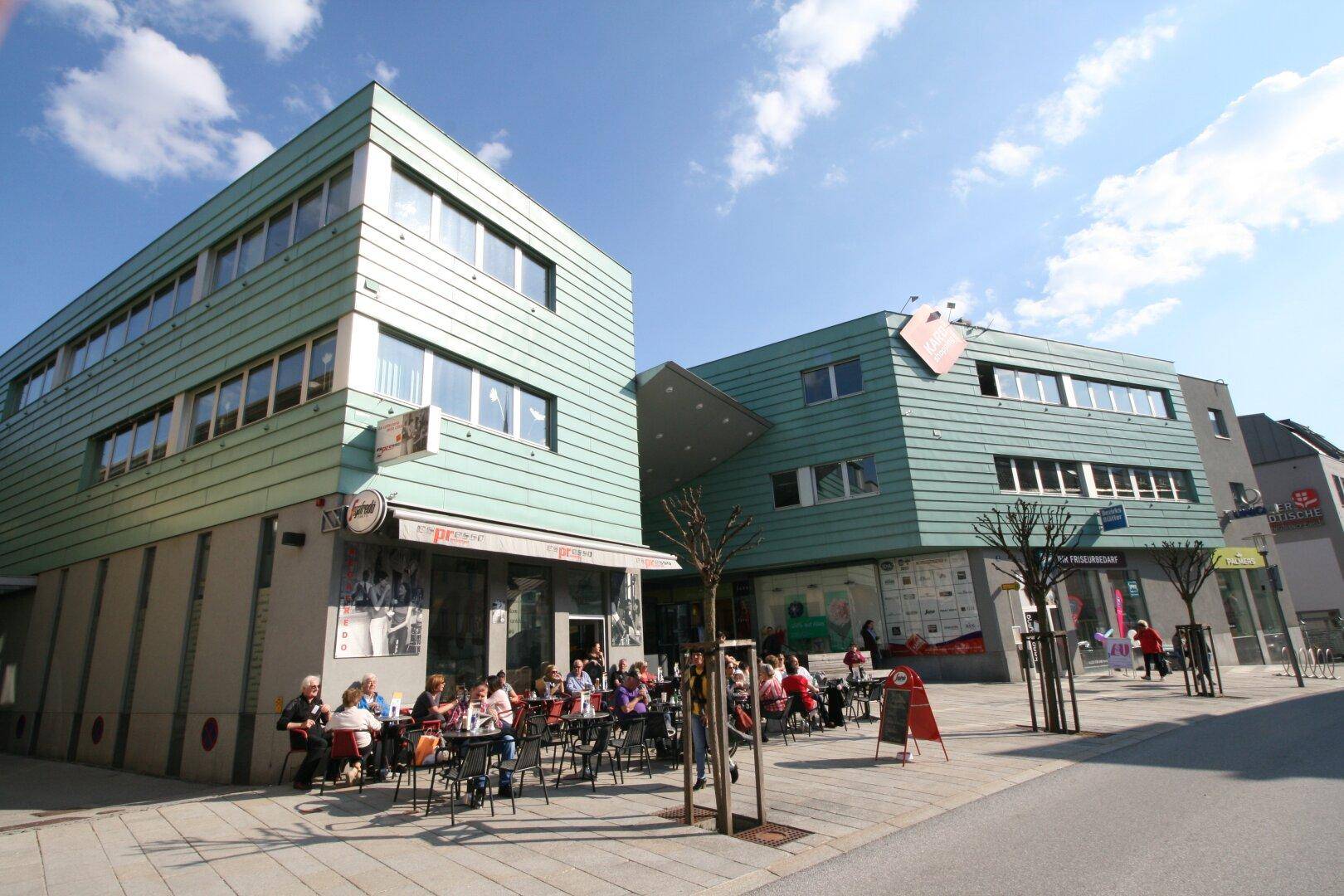 Moderne Gebäude mit grüner Fassade und einem belebten Café-Terrassenbereich im Freien.