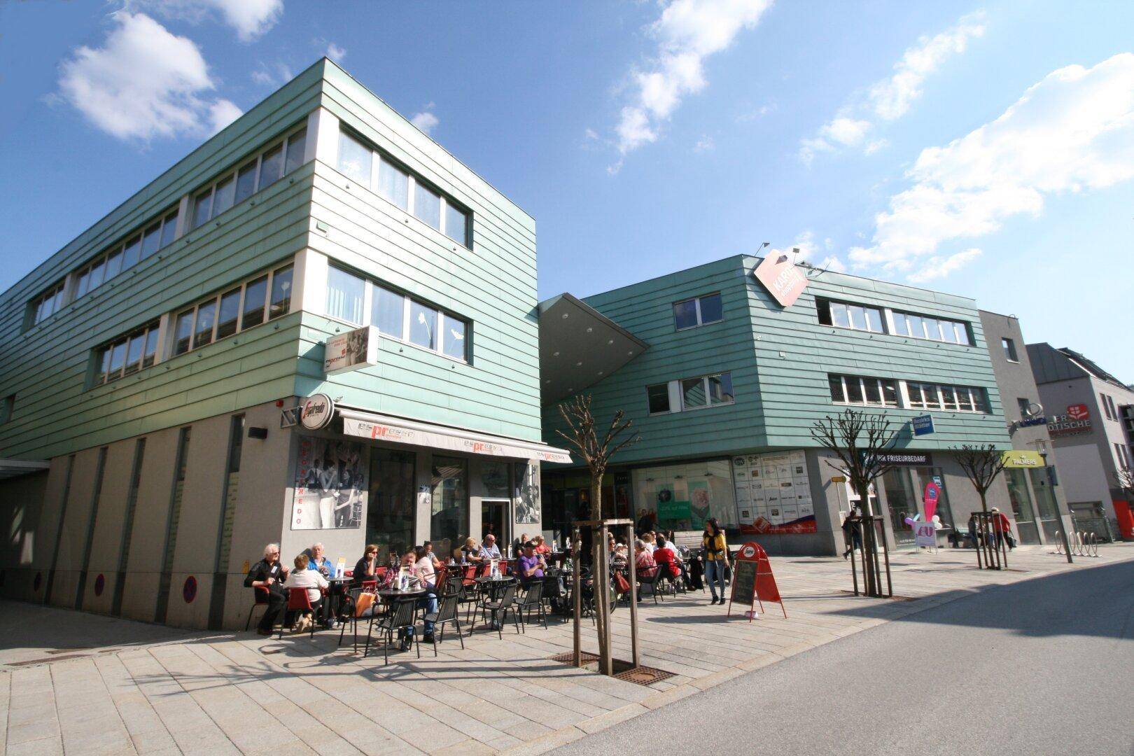 Moderne Gebäude mit grüner Fassade und einer belebten Café-Terrasse im Freien unter blauem Himmel.