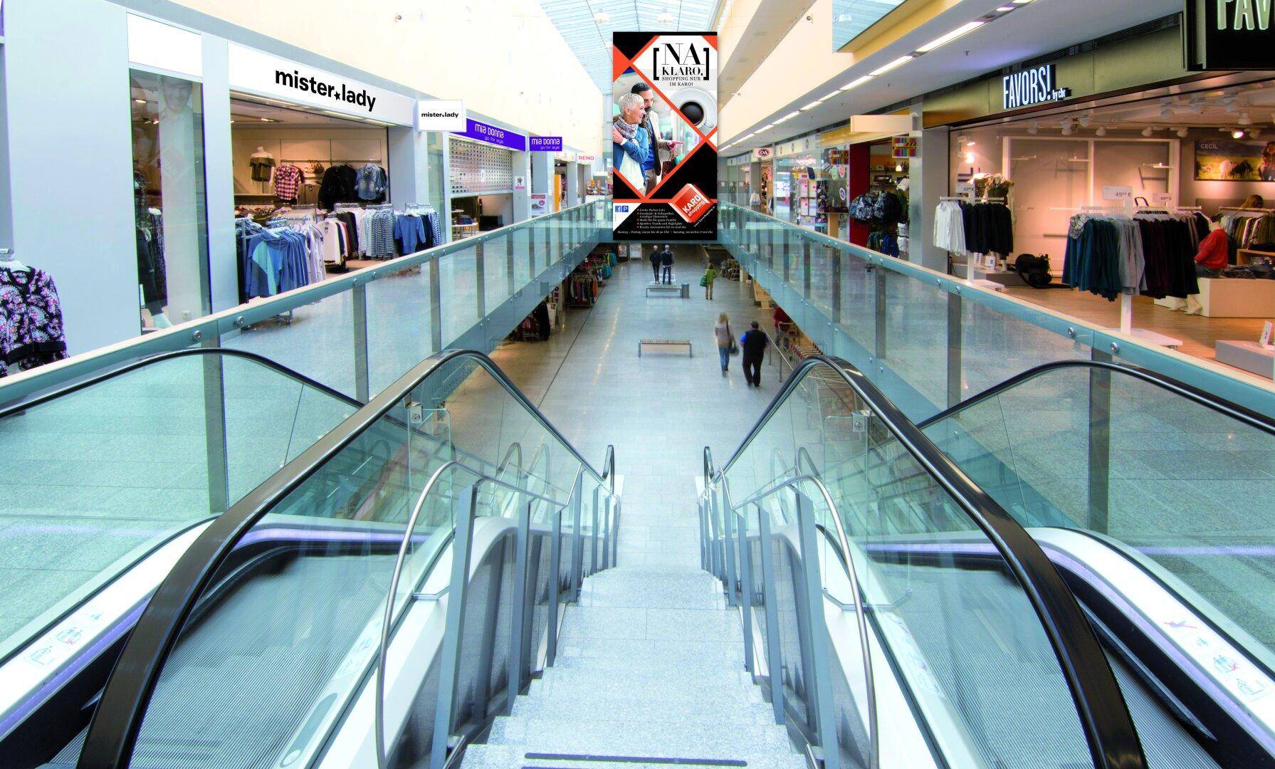 Blick von oben auf eine Rolltreppe in einem modernen Einkaufszentrum mit Geschäften und Glasgeländern.