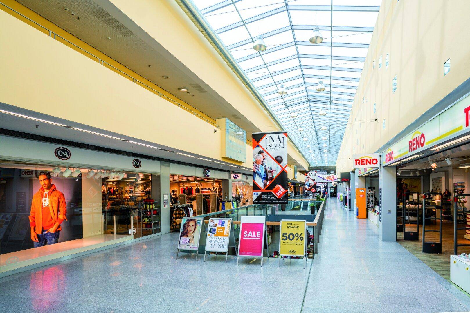 Langer Gang in einem Einkaufszentrum mit verschiedenen Geschäften, Werbetafeln und einem hellen Glasdach.