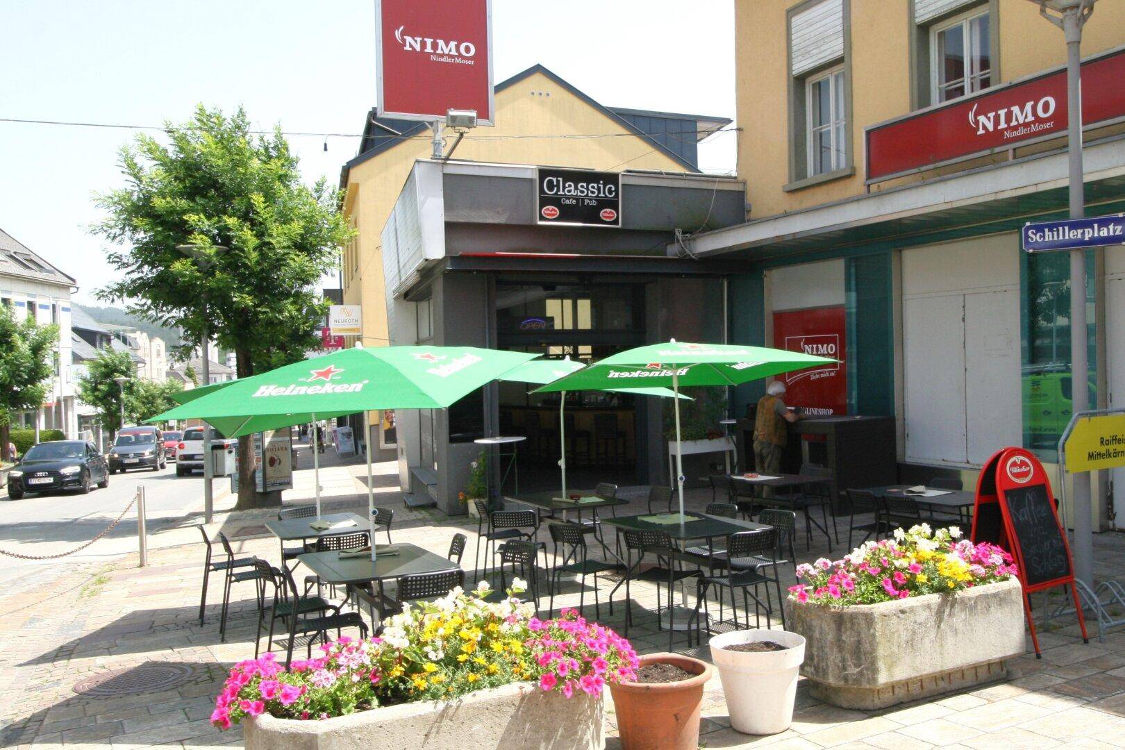 Außenbereich eines Cafés mit Tischen, Stühlen, grünen Sonnenschirmen und Blumenkästen an einer Straße.