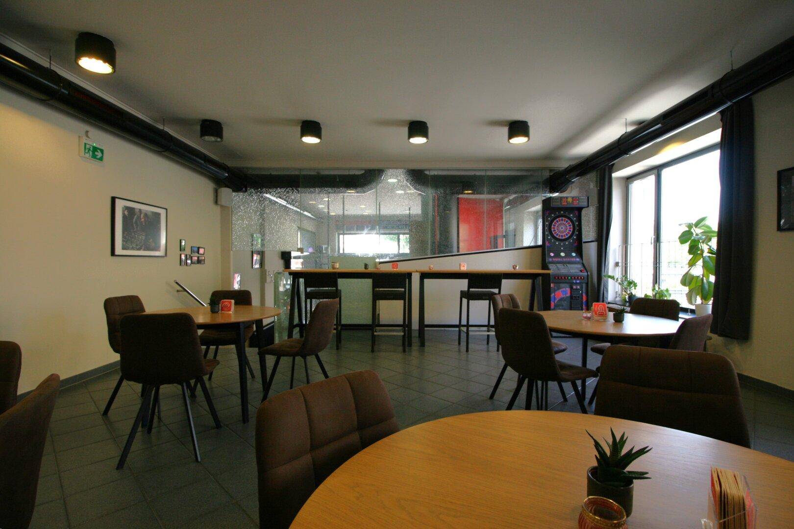 Gemütlicher Loungebereich mit runden Tischen, bequemen Stühlen und einem Dartboard im Hintergrund.