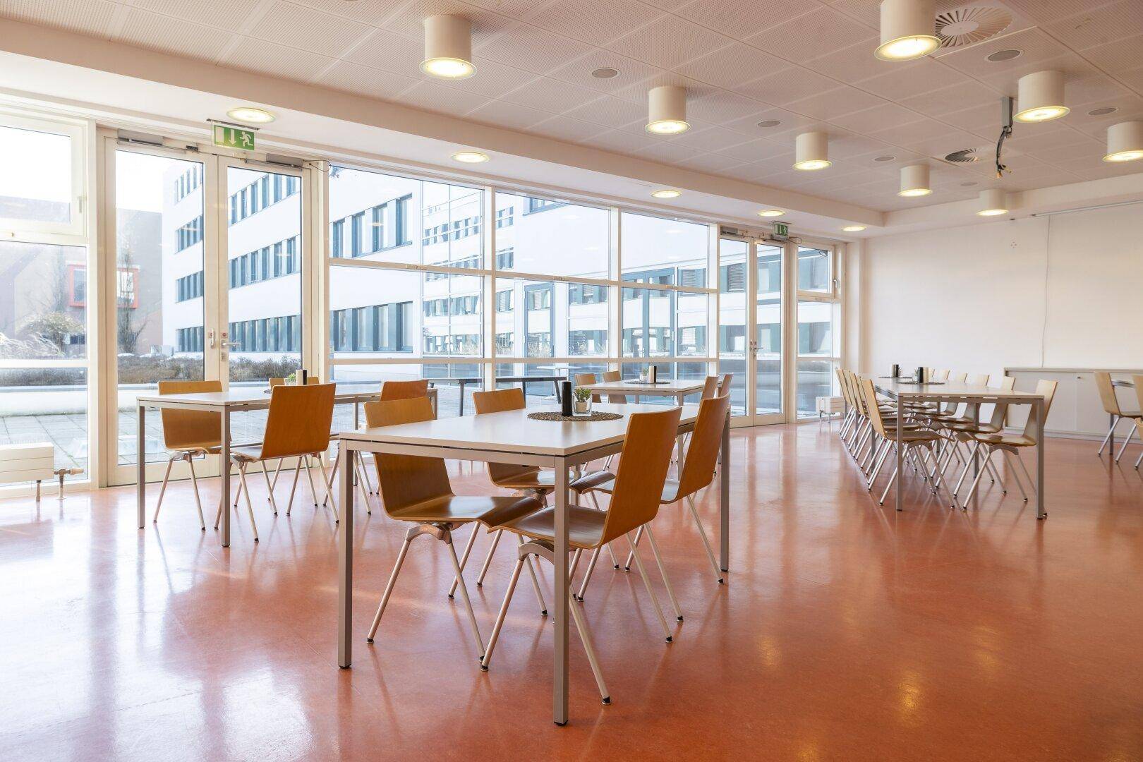 Geräumiger Speisesaal mit vielen Tischen, Stühlen und großen Fenstern mit Blick nach draußen.