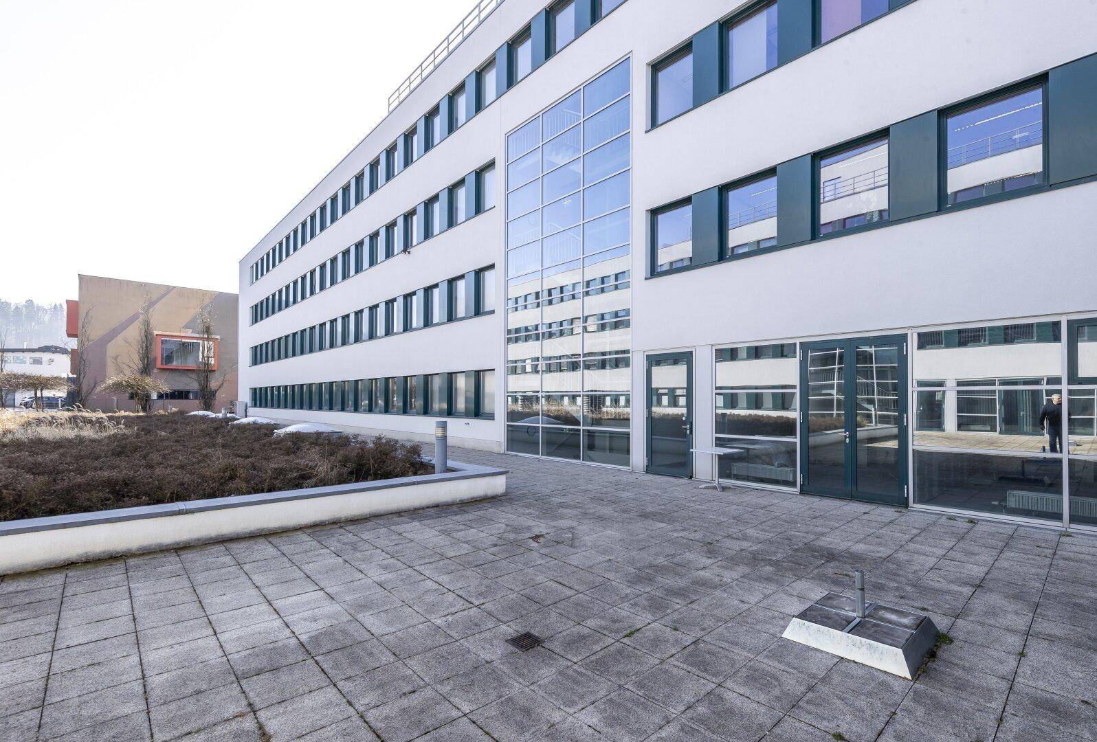 Moderne Gebäudefassade mit großen Glasflächen und einer gepflasterten Terrasse im Vordergrund.