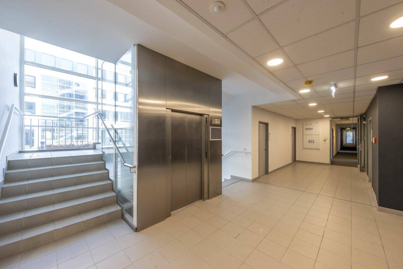 Heller Flur mit Aufzug und Treppenhaus, ausgestattet mit Fliesenboden und großen Fenstern für viel Tageslicht.