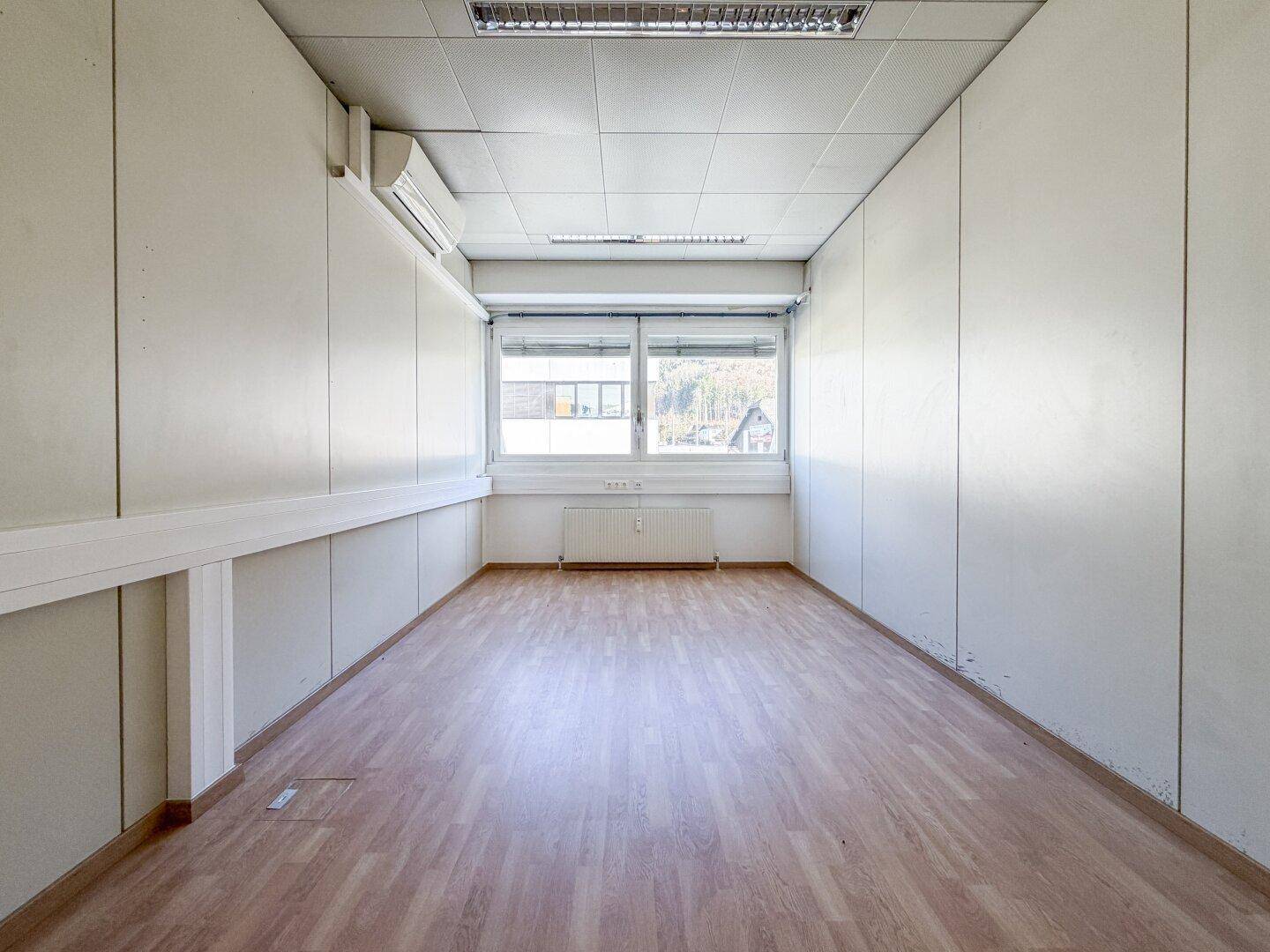 Schmaler Büroraum mit Laminatboden, Klimaanlage und einem Fenster, das Tageslicht hereinlässt.