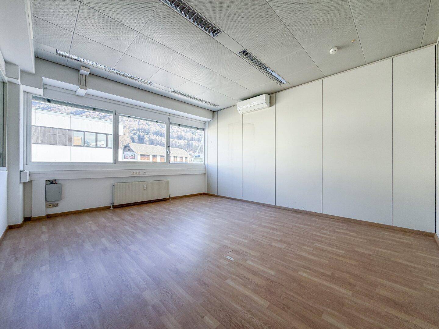 Leerer Büroraum mit Laminatboden, Klimaanlage und Fenstern, die einen Blick auf die Umgebung bieten.