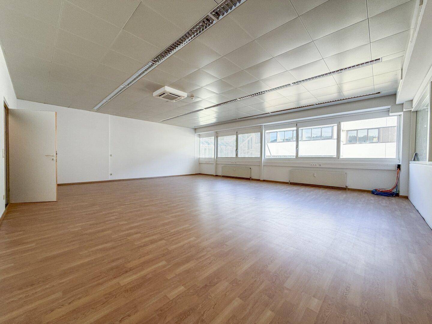 Heller Büroraum mit Laminatboden, abgehängter Decke und großen Fenstern, die viel Tageslicht hereinlassen.