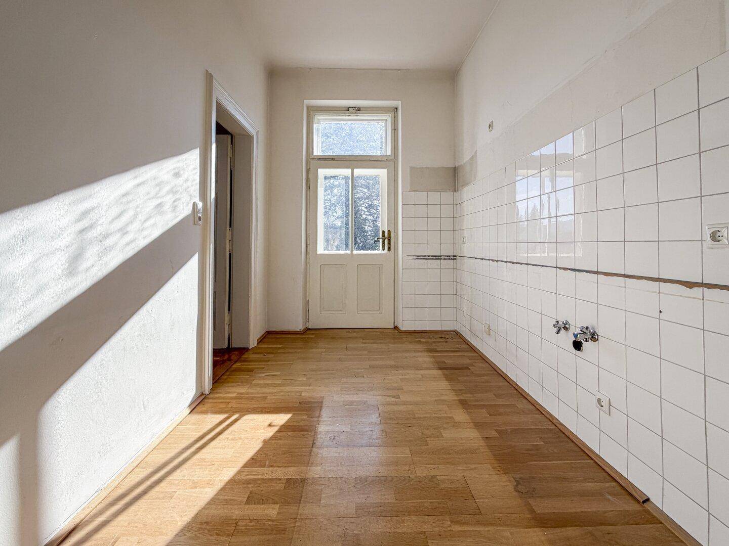 Leere Küche mit Holzboden, weißen Wandfliesen und einer Tür, die zu einem Außenbereich führt.
