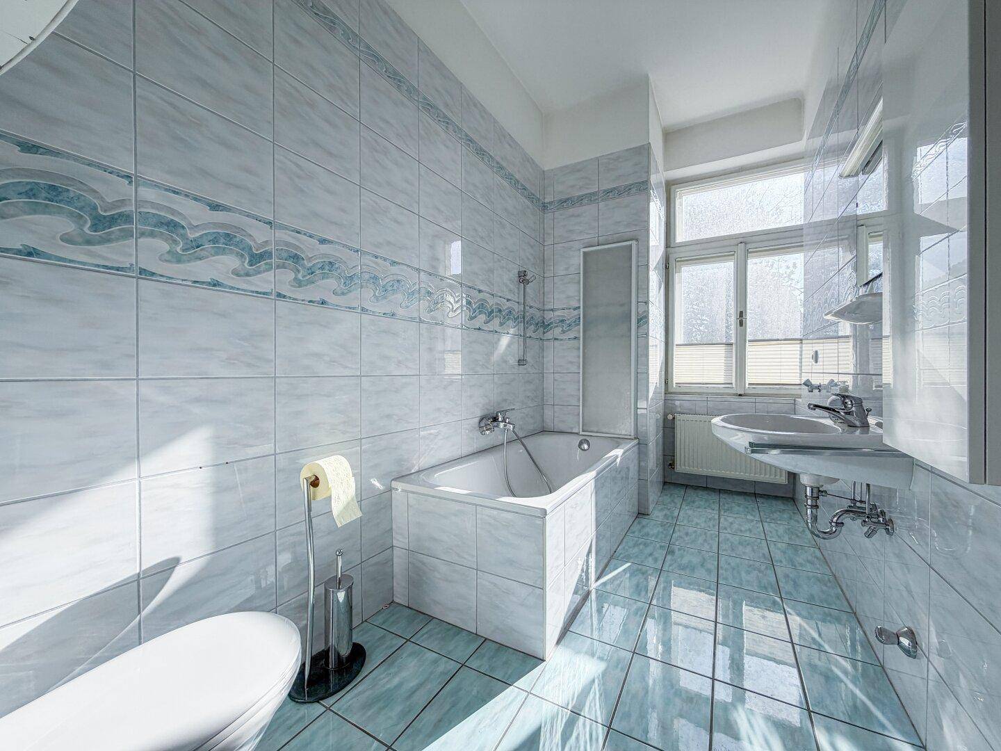 Badezimmer mit hellblauen Fliesen, Badewanne, Waschbecken und Fenster für natürliches Licht.