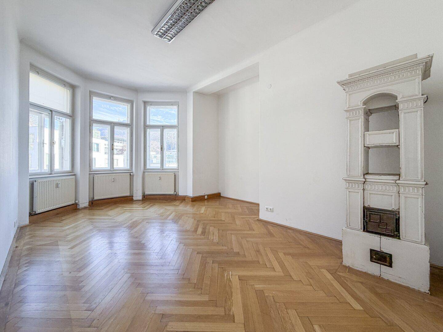 Heller Raum mit Fischgrätparkett, einem dekorativen Kachelofen und großen Erkerfenstern.
