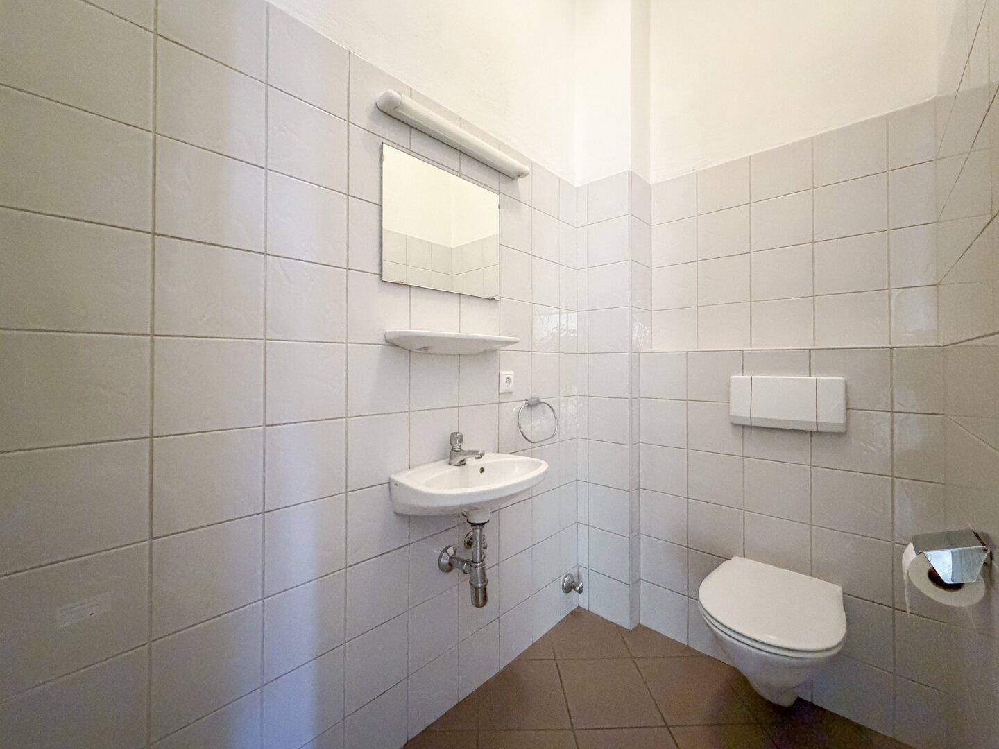 Kleines Badezimmer mit weißen Wandfliesen, Waschbecken und wandhängender Toilette.