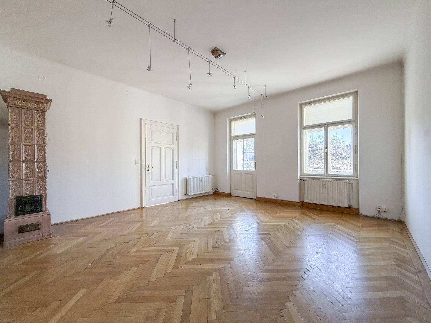 Geräumiges Zimmer mit Fischgrätparkett, einem traditionellen Kachelofen und großen Fenstern.