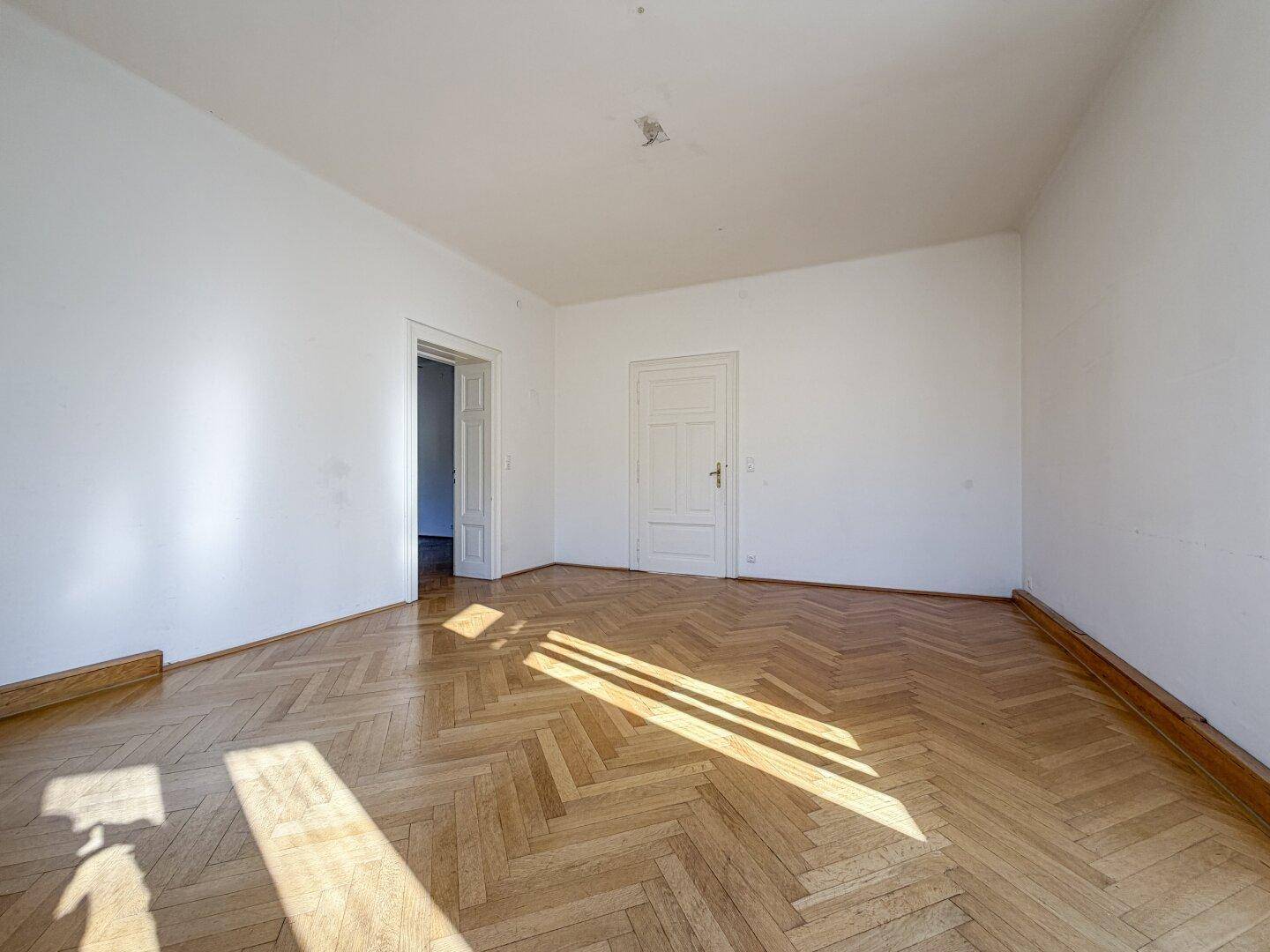 Helles, leeres Zimmer mit Fischgrätparkett und einer klassischen Tür, durch die viel Sonnenlicht fällt.