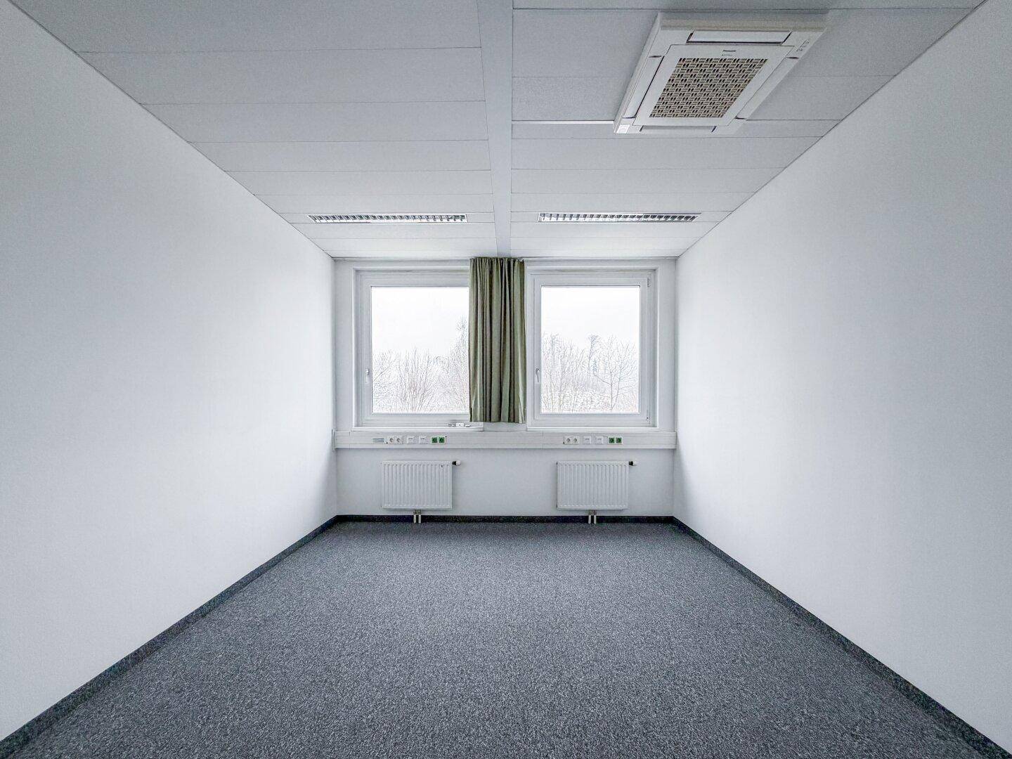 Kompakter Büroraum mit Teppichboden, zwei Fenstern und Klimaanlage, ideal für ein Einzelbüro.
