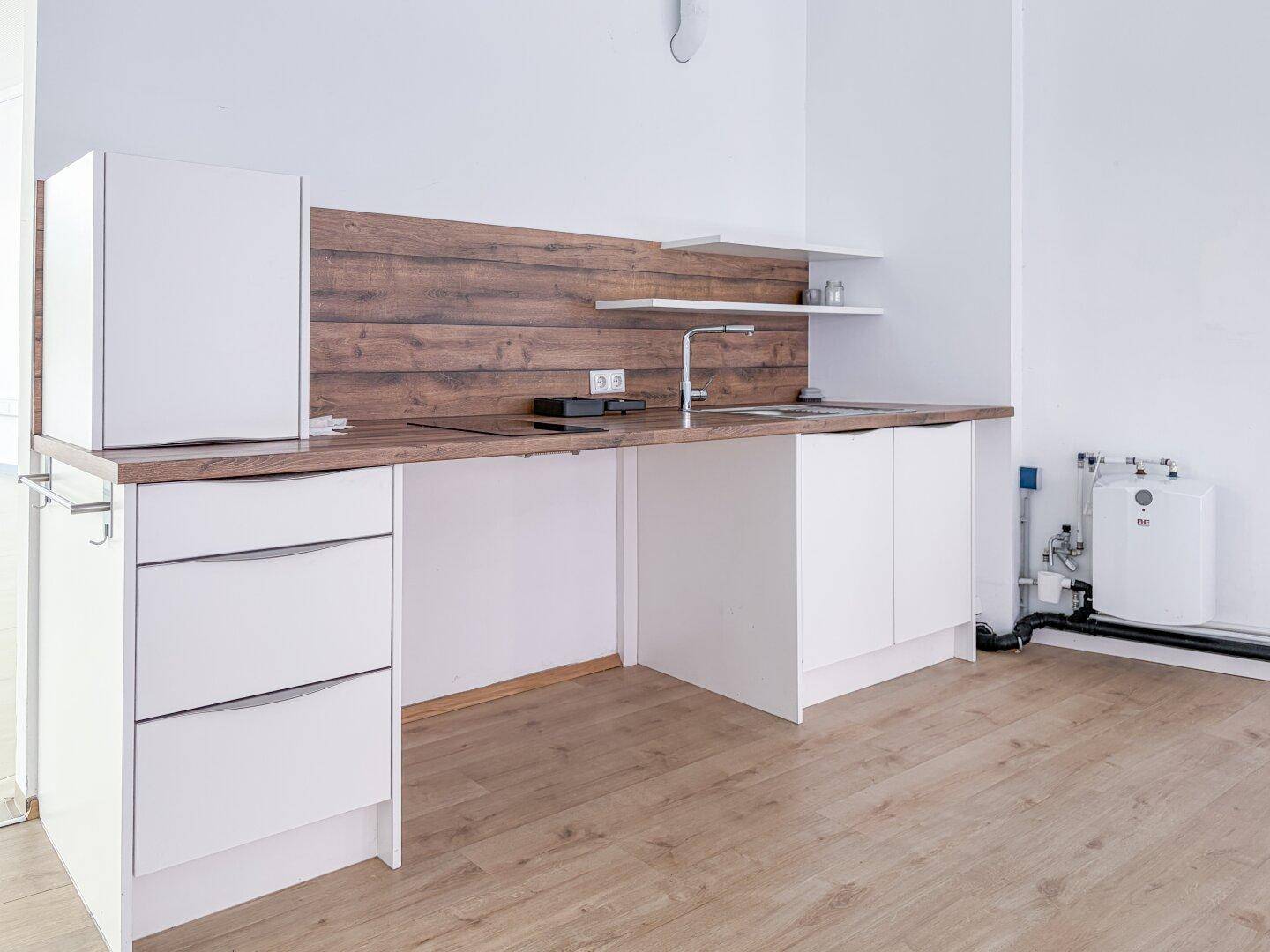 Kleine Küchenzeile mit weißen Schränken, Holzrückwand, Spüle und Kühlschrank, ideal für Pausenbereiche.