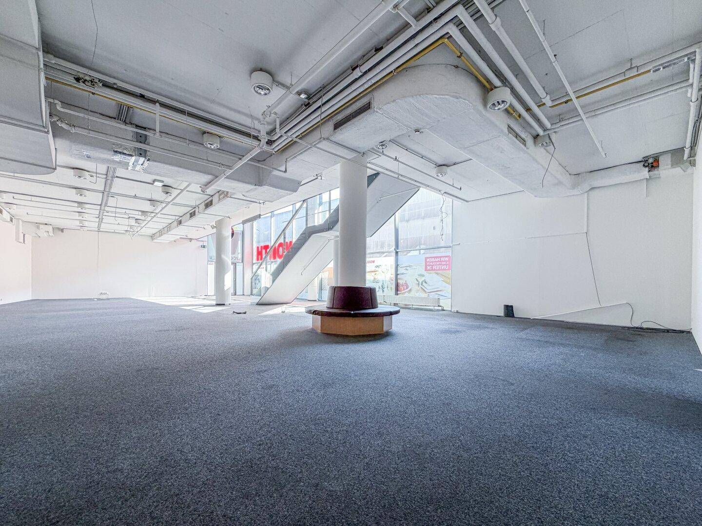 Großer, leerer Verkaufsraum mit Teppichboden, runder Sitzgelegenheit und sichtbaren Deckeninstallationen.