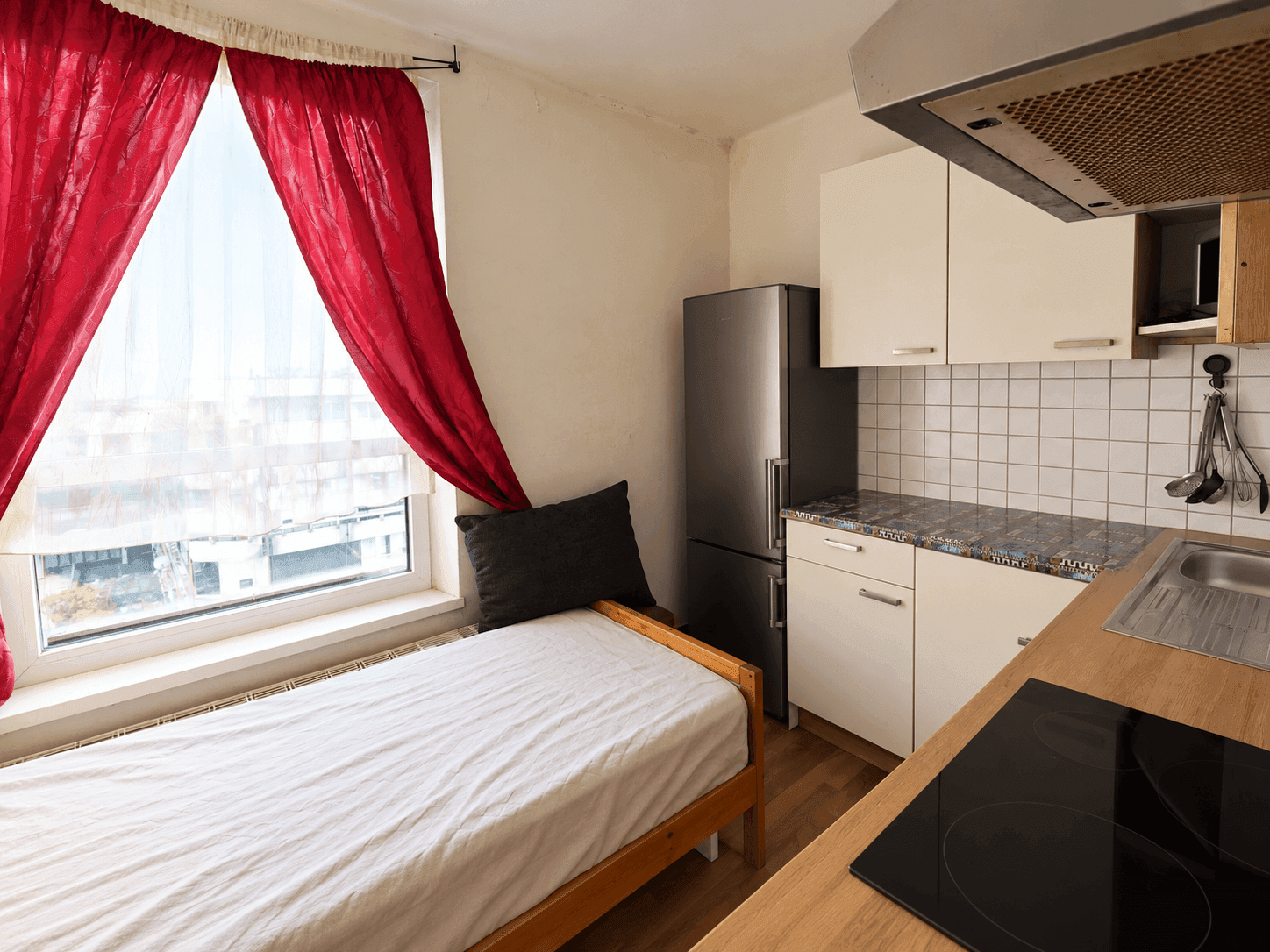 Kleine Küche mit Einzelbett, roten Vorhängen und Blick auf die umliegenden Gebäude.