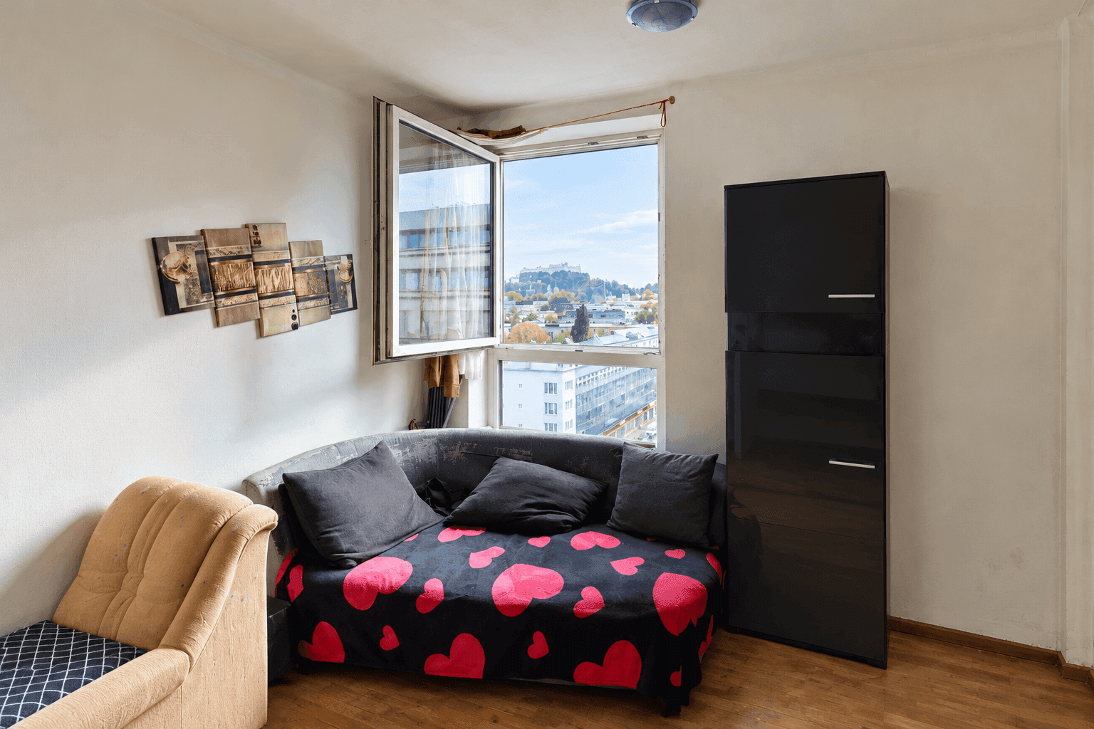 Wohnzimmer mit Sofa, schwarzem Schrank und einem Fenster mit Blick auf die Stadt.