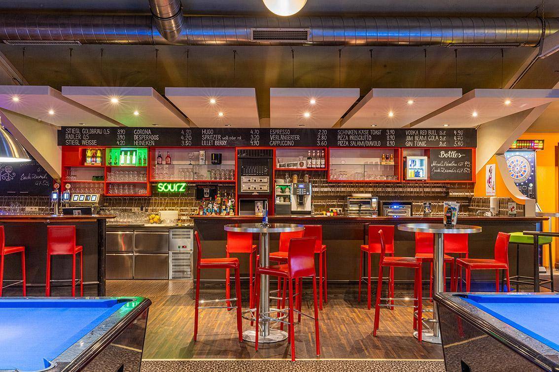 Lebhafte Bar mit roten Hockern, einer großen Theke und Billardtischen im Vordergrund.