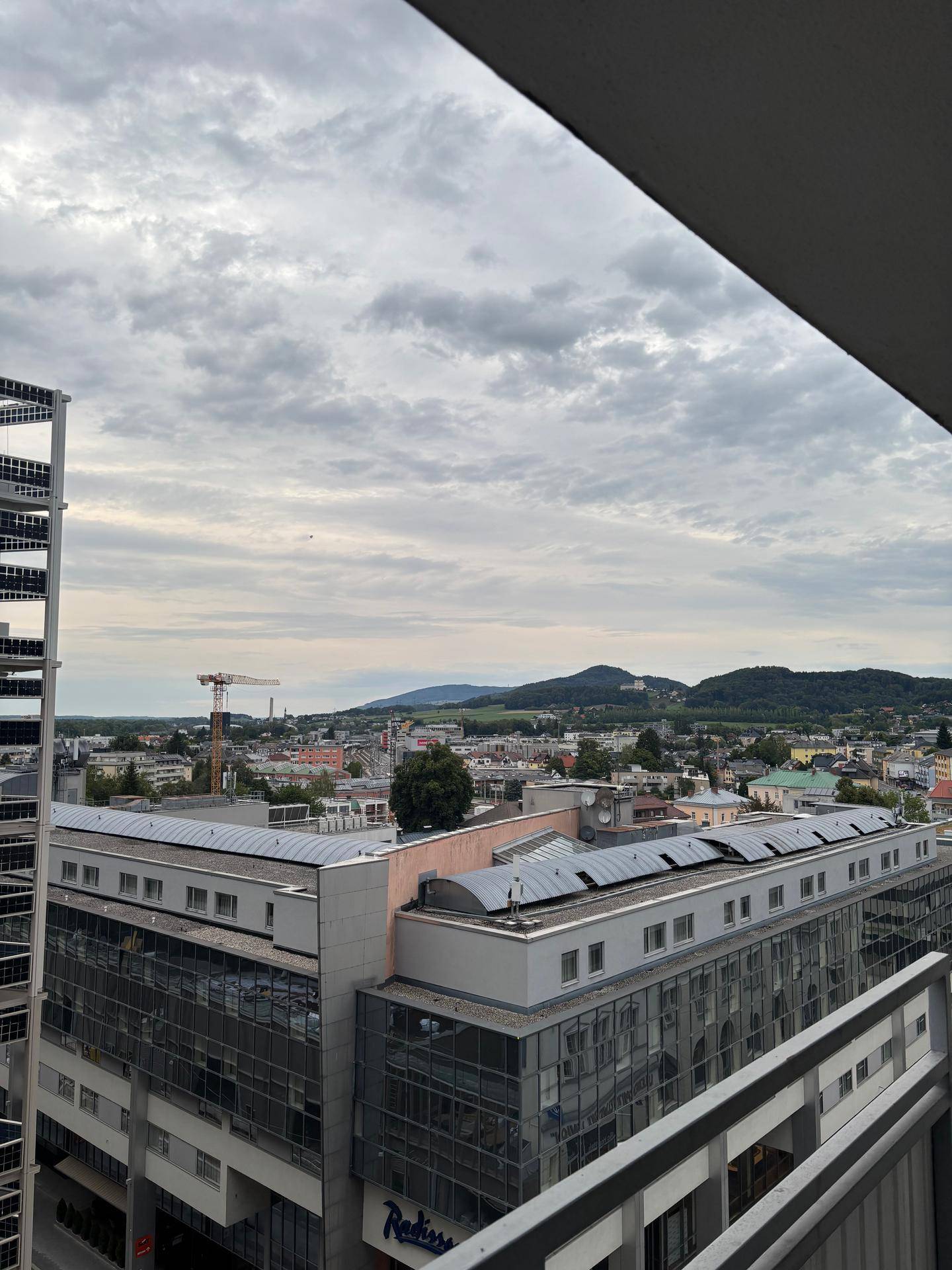 Weitblick vom Balkon über die Stadtlandschaft mit Bergen im Hintergrund unter bewölktem Himmel.