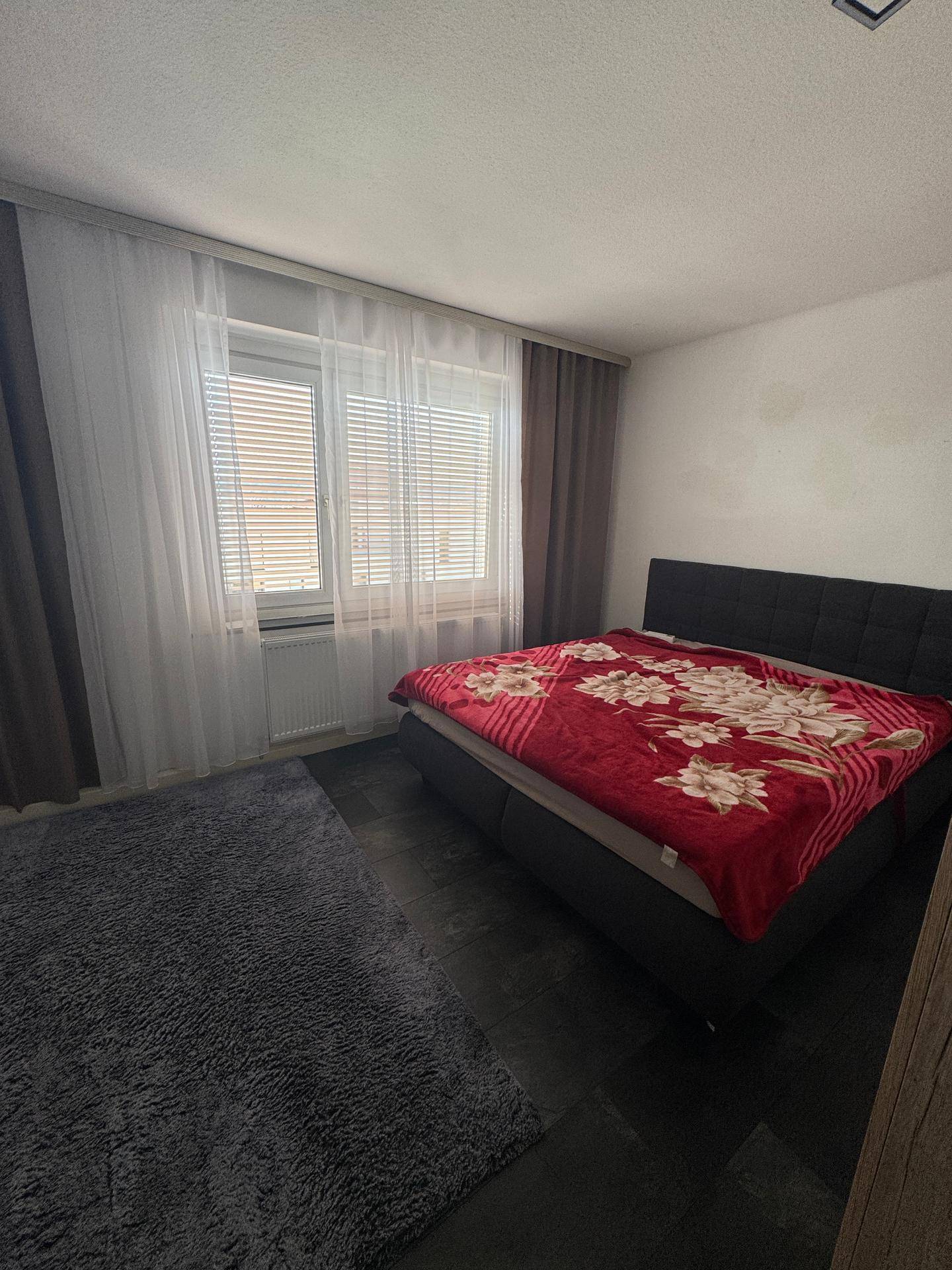 Helles Schlafzimmer mit Doppelbett, Teppich und Fenster mit Jalousien.