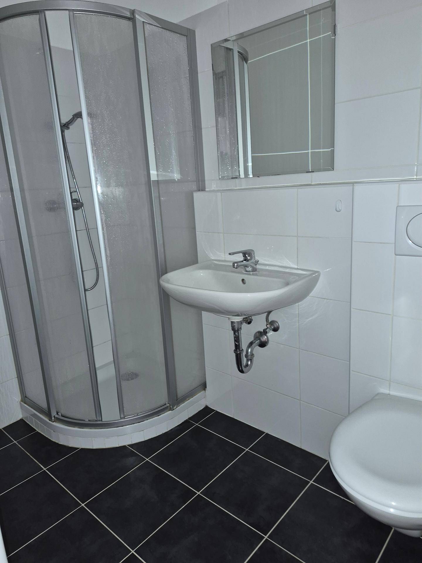 Badezimmer mit Duschkabine, Waschbecken, Wand-WC und dunklen Bodenfliesen in modernem Design.