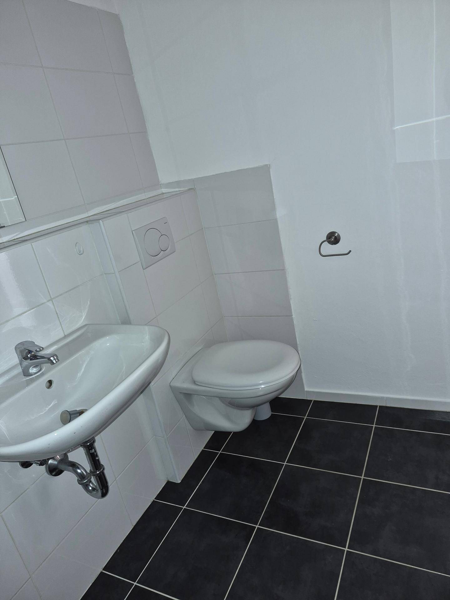 Kleines Gäste-WC mit Wand-WC, Waschbecken und dunklen Bodenfliesen in sauberem Zustand.