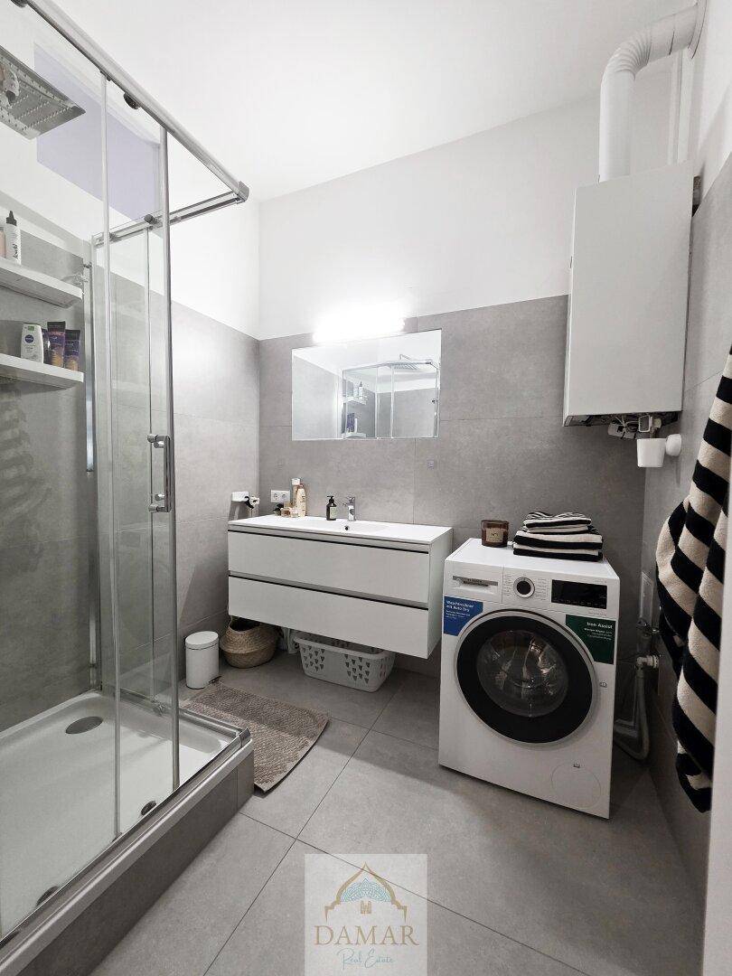 Modernes Badezimmer mit Duschkabine, Waschbecken und Waschmaschine, ausgestattet mit grauen Fliesen.